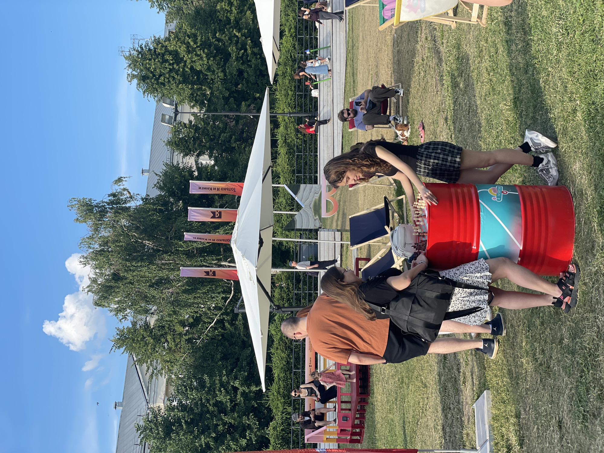 The image depicts an outdoor scene in Kazan, Russia, possibly during a festival or event. In the foreground, two young women are standing next to a large red barrel which is being used as a table for a chess game. One woman, wearing a black tank top and a plaid skirt, is leaning on the barrel, looking at the chess pieces. The other woman, in a black top and a white patterned skirt, stands beside her, her hand on the barrel as well. Behind them, a man in an orange t-shirt and black shorts is walking away from the camera.
Further back, several other individuals are visible. A family is seated on a colorful bench structure, with a young girl in a pink dress dancing in front of them. Other people are scattered around the grassy area, some sitting on lounge chairs and others walking. In the background, there are large white umbrellas providing shade, trees, and a building with a gray roof. Banners with Russian text hang from poles. The overall atmosphere appears to be relaxed and festive, with people enjoying a sunny day outdoors.