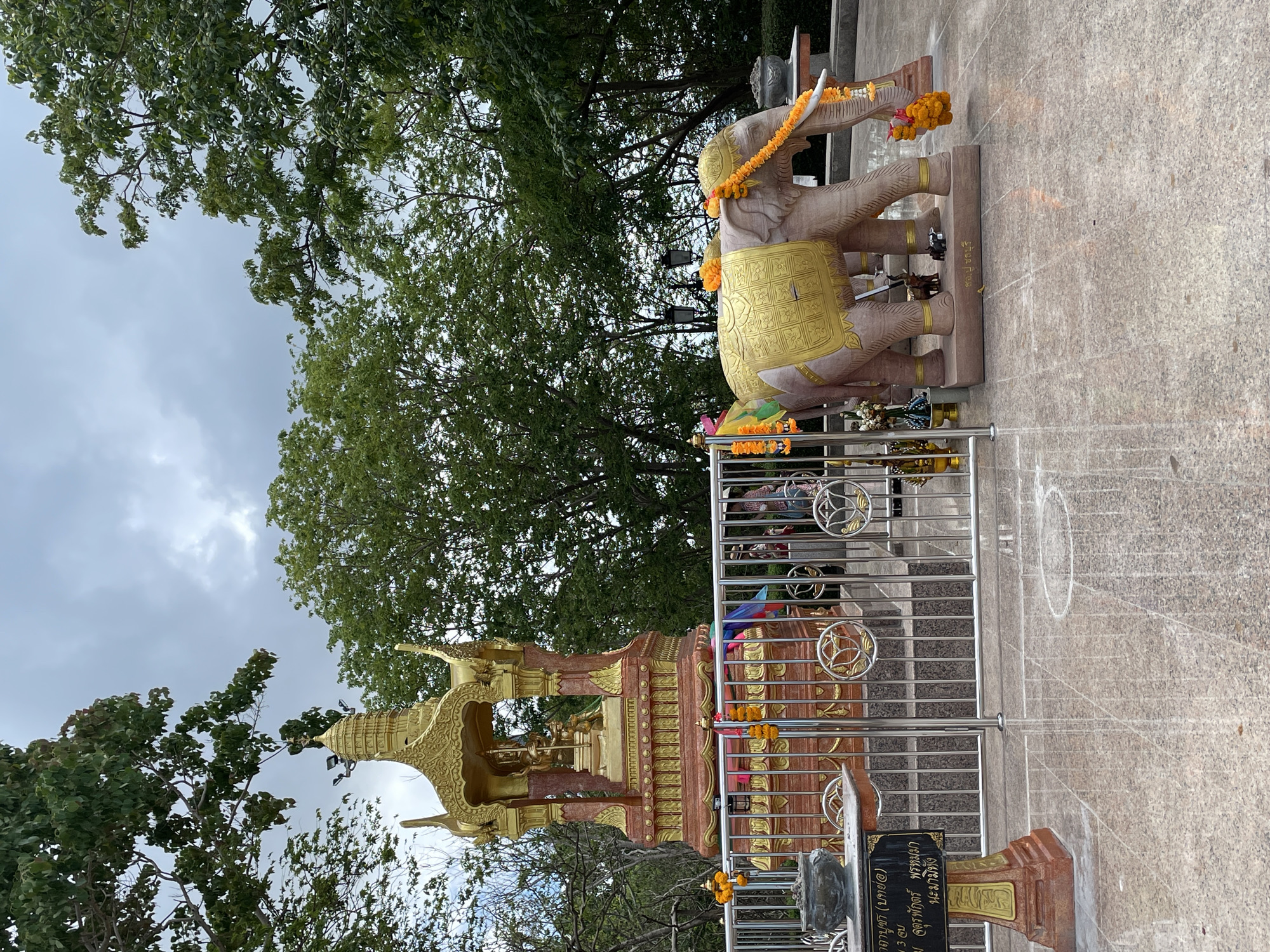 This outdoor image captures a religious site in Rawai, Thailand, featuring two prominent structures: a statue of a pair of elephants and a golden shrine. The elephant statue is draped with garlands and appears to be made of stone, with a golden cloth covering its back. It stands on a concrete surface. A metal railing surrounds the area, with additional decorations and offerings visible. The golden shrine is intricately designed and adorned with multiple tiers and decorative elements, also surrounded by the same metal railing. A sign with Thai script is placed near the shrine. Lush green trees and a cloudy sky form the backdrop of the scene.