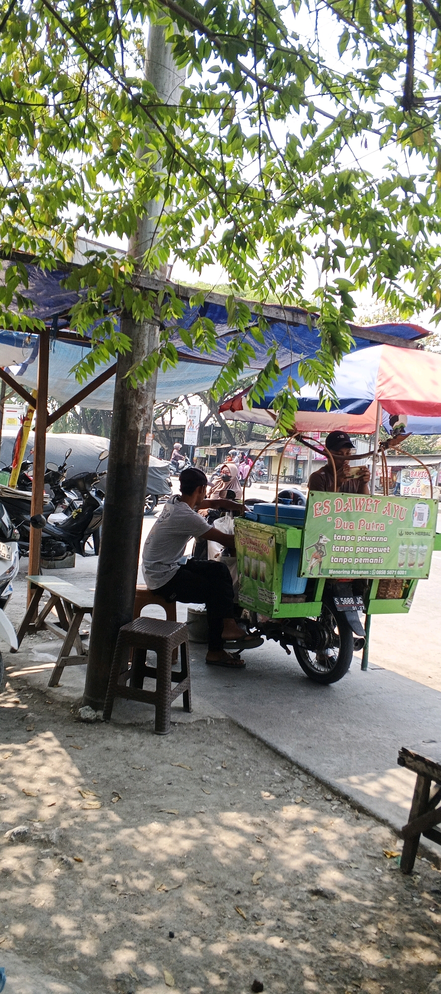 In Sukodadi, Indonesia, a street vendor operates a mobile ice cart. The cart, a bright green box mounted on a motorcycle, displays advertisements for "ES DAWAET AYU" and "Dua Putra," highlighting that the product is made without artificial coloring, preservatives, or flavorings. The cart also features images of three drinks. Two men are present: one, wearing a grey t-shirt and black pants, sits on a small brown plastic stool, facing away from the camera and towards the cart, appearing to be engaged in preparing or serving the product. The second man, wearing a dark t-shirt and a black cap, sits on the cart's rear, holding a mobile phone to his ear, possibly taking an order or making a call.
The scene is set outdoors, under the dappled shade of a tree with abundant green leaves. Behind the cart, a makeshift stall with a blue tarp roof is visible, supported by wooden poles. Several motorcycles are parked nearby, suggesting a busy roadside location. The ground is a mix of dirt and concrete, with scattered dry leaves. Sunlight filters through the leaves, creating a pattern of light and shadow on the ground. In the background, other vendors and people on motorcycles are visible, indicating a typical street scene in a commercial area. The overall atmosphere suggests a typical day at a roadside business in Indonesia.