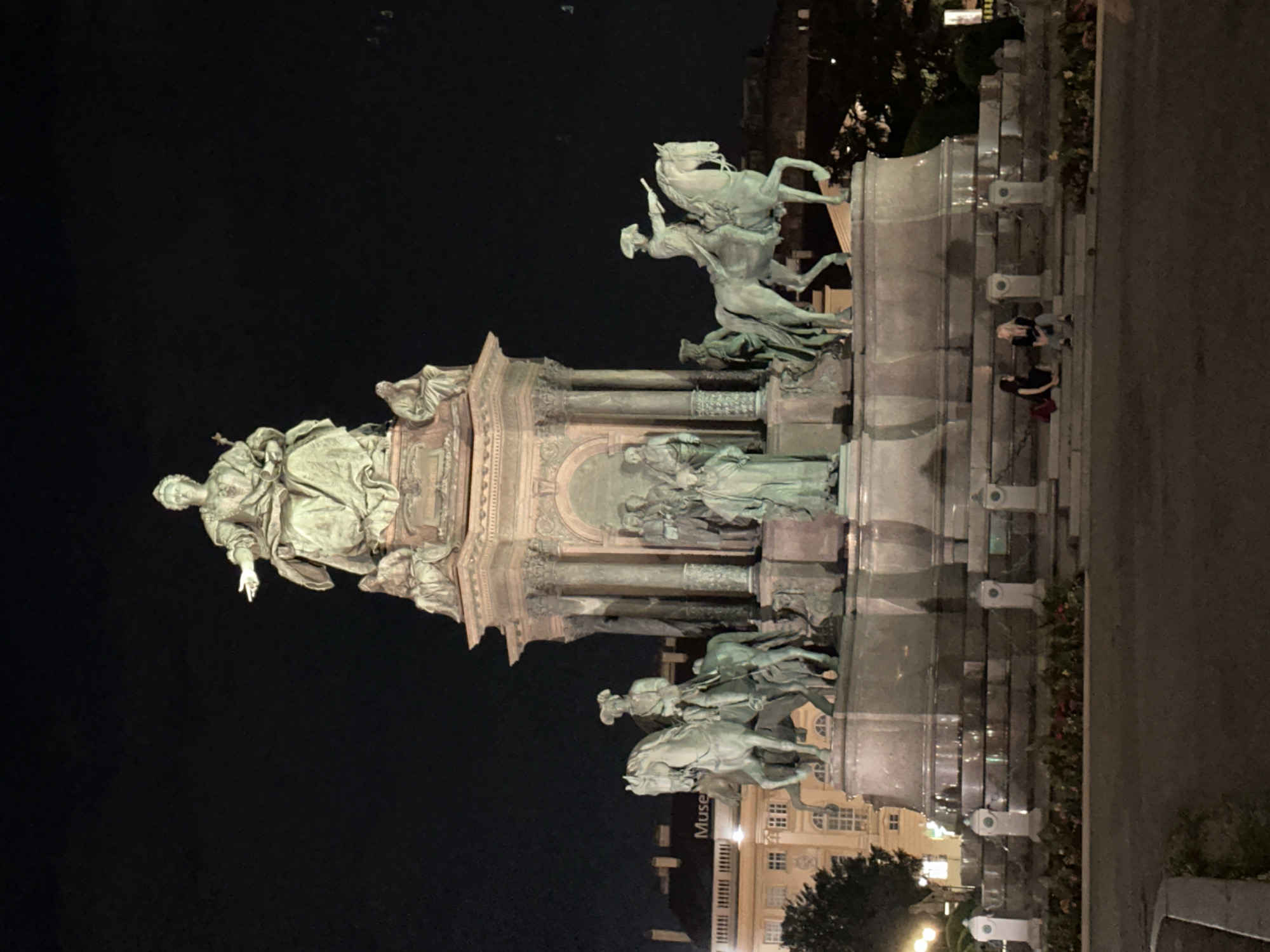 The image shows a nighttime view of the Maria Theresien Monument in Vienna, Austria. The monument is a large, ornate structure featuring statues of Empress Maria Theresa and several other figures, including equestrian statues. The monument is illuminated, and the surrounding area appears to be a public space, possibly a park or plaza. In the foreground, two individuals are seated on the steps of the monument. One is wearing a black dress, and the other is wearing a jacket and pants. They appear to be interacting or conversing. Buildings are visible in the background, some of which are illuminated. The overall impression is of a historical and architectural landmark at night.