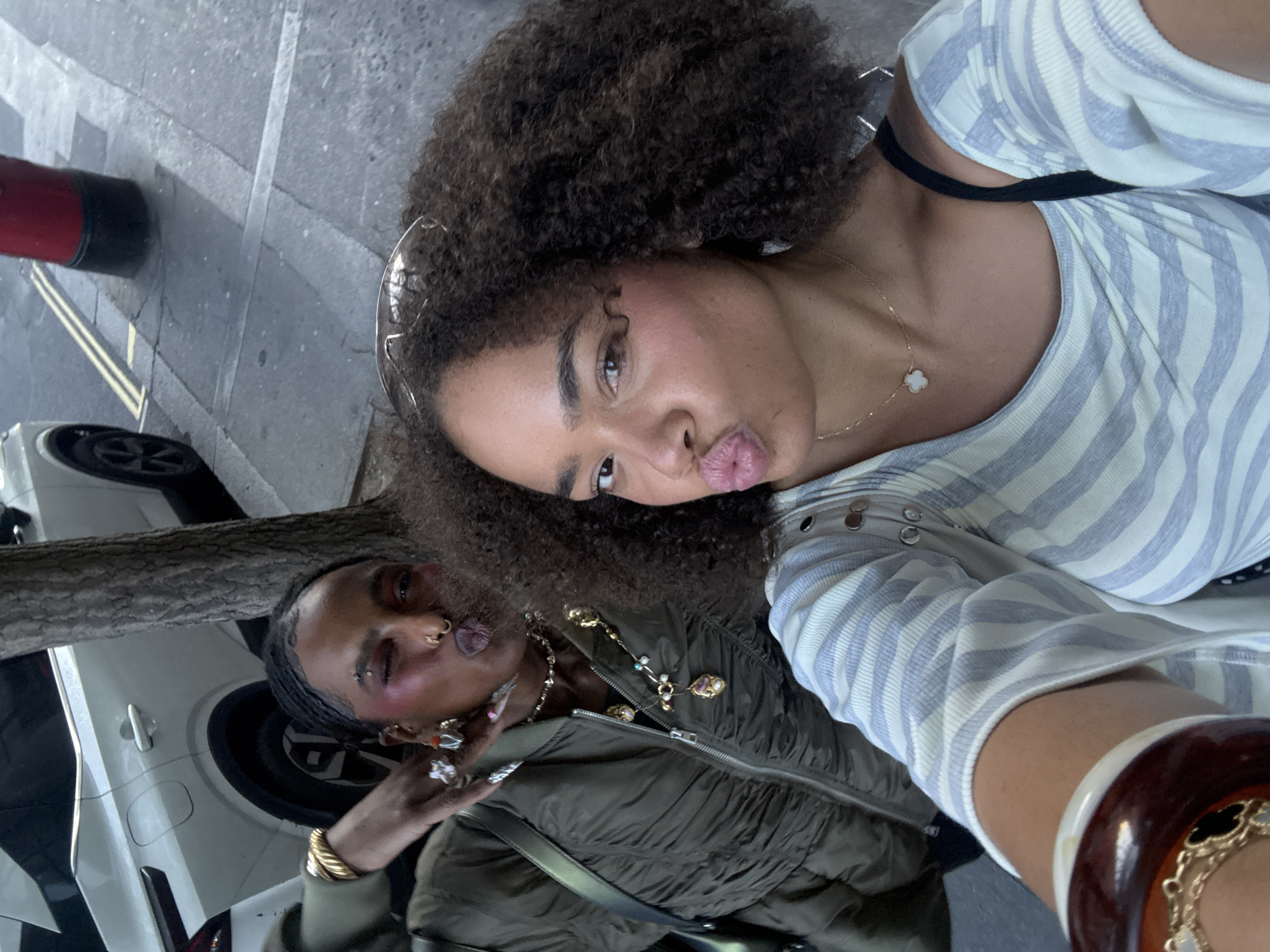 Two individuals pose for a selfie in what appears to be a city setting. The person in the foreground, presumably holding the camera, has curly hair and is wearing a striped top with a black undergarment strap visible. They are making a pouting expression. The second person, positioned slightly behind and to the side, is wearing an olive green jacket, adorned with multiple necklaces and rings, and also makes a playful expression, sticking out their tongue. The background includes a section of a car, a tree trunk, and what looks like a red and black bollard next to a curb and street surface, suggesting an urban environment. The overall scene is captured with an upward angle.
