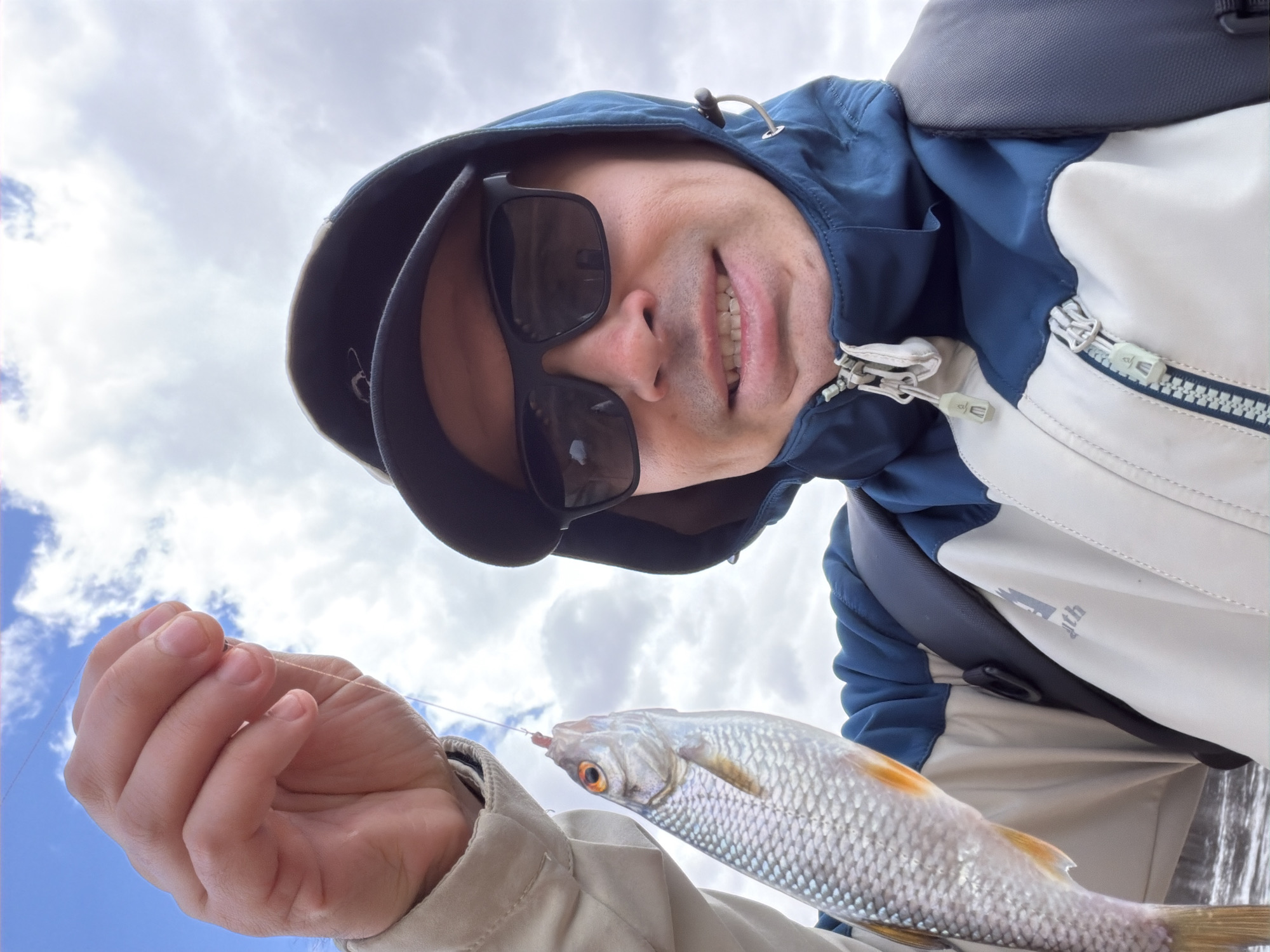 A man is holding a fish up to the camera. He is wearing a black baseball cap, sunglasses, and a blue and white jacket. The sky is cloudy. There is no identifiable location in Izhevsk, Russia visible.