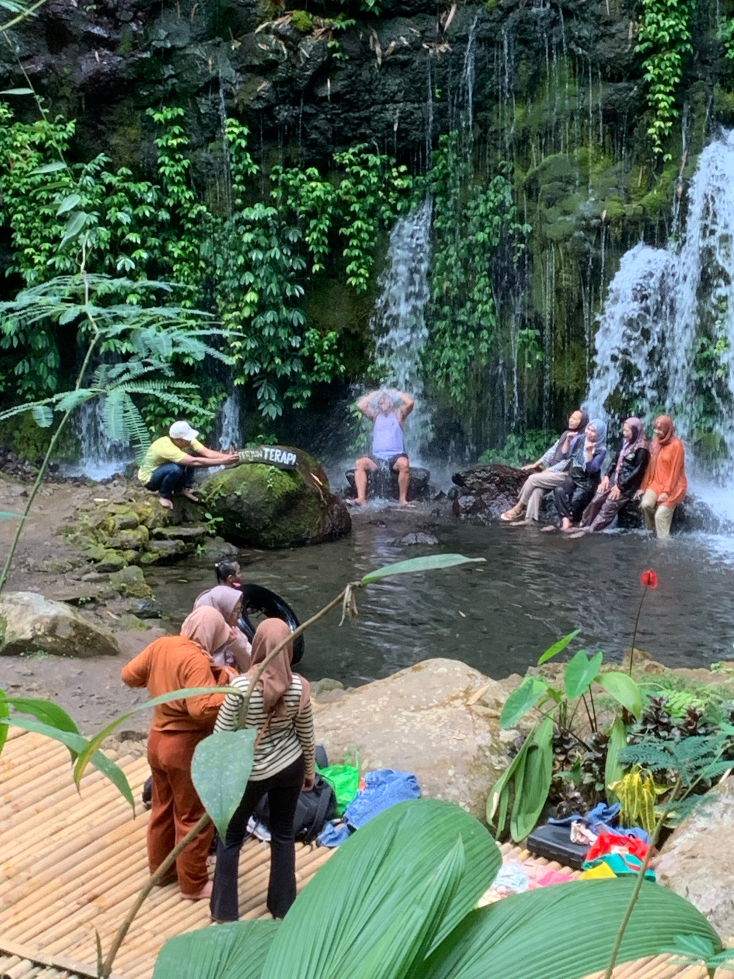 In Pronojiwo, Indonesia, a group of people are enjoying a waterfall and a natural pool. One man in a yellow shirt and white cap is crouched by a large moss-covered rock, gesturing towards a sign on the rock. A man in a blue tank top and white shorts is standing in the waterfall, with water cascading over him. To his right, five people, three women and two others, are sitting on rocks in the pool, with water flowing around them. In the foreground, three people, two women and one person whose gender is not discernible, are standing on a bamboo platform. One woman in an orange shirt and brown pants is facing away from the camera, while another woman in a striped shirt and black pants is also facing away. A third person is partially visible between them. Various items, including bags and clothing, are scattered on the bamboo platform and nearby rocks. The environment is lush with green vegetation covering the rocks and surrounding the waterfall.