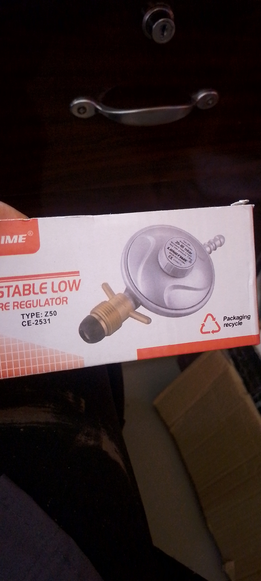 A close-up, low-angle shot features a box for a "LOMTIME ADJUSTABLE LOW PRESSURE REGULATOR". The box is white with red accents. Text on the box reads "LOMTIME", "ADJUSTABLE LOW PRESSURE REGULATOR", "TYPE: Z50", and "CE-2531". A silver regulator with brass fittings is visible on the right side of the box, along with a recycling symbol and the words "Packaging recycle". The box appears to be placed on a dark surface, possibly a drawer, with a metal handle and lock visible at the top of the frame. The lower portion of the image shows a person's dark clothing and a corrugated cardboard box. The scene is indoors, with no specific identifiable landmarks to determine a location within Jalingo, Nigeria.