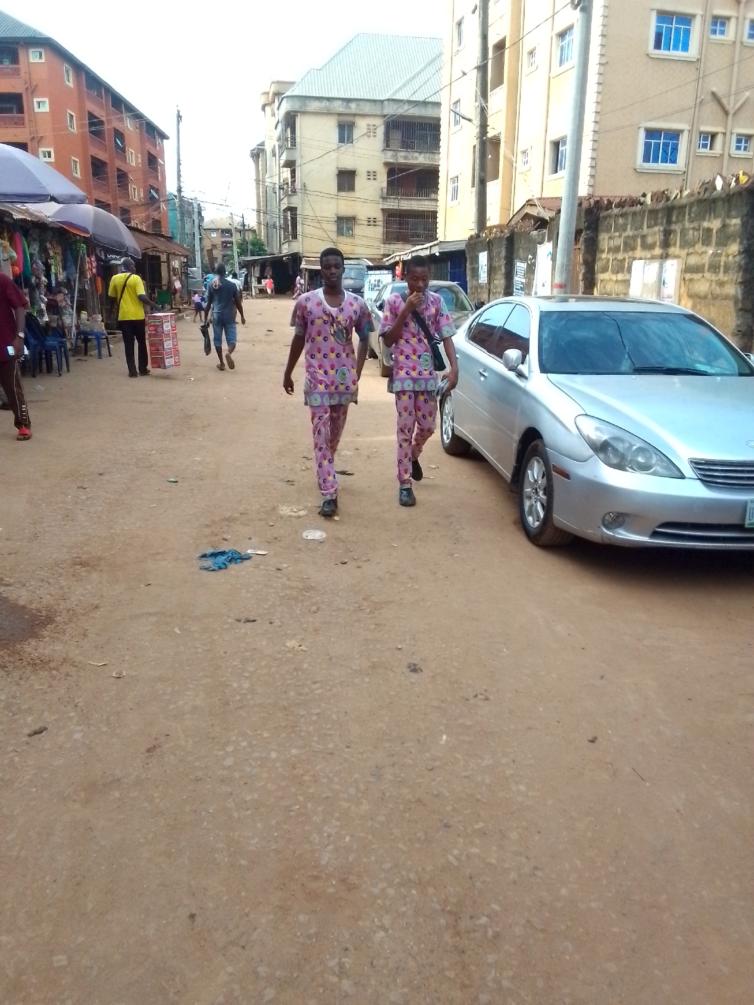 The image shows a street in Onitsha, Nigeria. Two young men wearing matching pink and purple patterned outfits are walking towards the camera on a dirt road. A silver car is parked on the right side of the road. Several buildings of varying heights line the street. People can be seen walking in the background, some near market stalls shaded by umbrellas. The environment appears dusty and somewhat crowded.