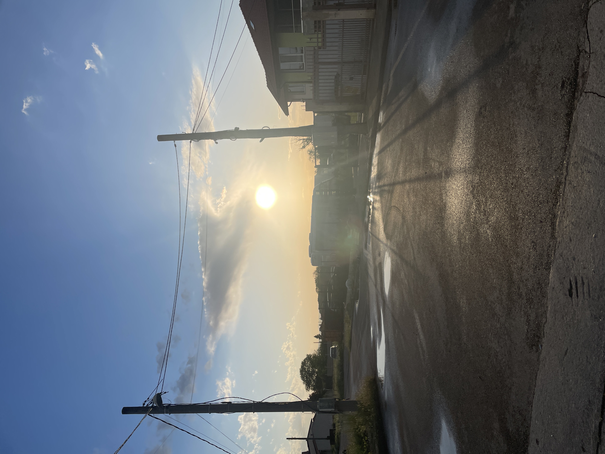 The image depicts a street scene in Sofia, Bulgaria during sunset. The street is wet and reflects the sunlight. A row of buildings is visible on the right side of the street. Utility poles and power lines run along the left side of the street, silhouetted against the sky. The sky is a clear blue transitioning to yellow near the sun. Clouds are present in the sky. There are puddles on the street. The image is oriented vertically.