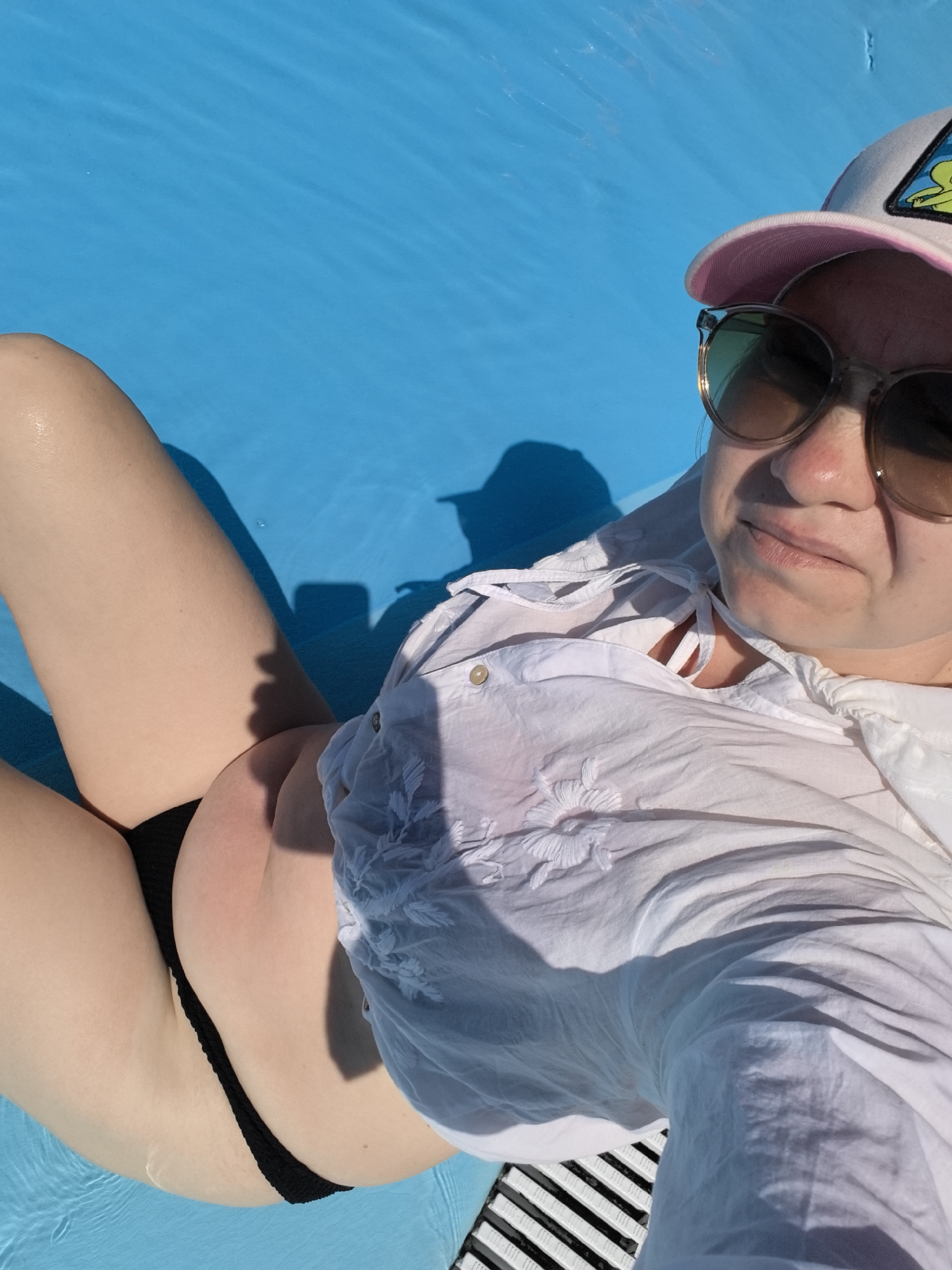 The image shows a person in black bikini bottoms and a wet white shirt with embroidery, lying on a bright blue surface that appears to be water, possibly a swimming pool. They are wearing a pink and white baseball cap and sunglasses, and their face is partially visible, looking towards the camera with a slight smile. Sunlight casts shadows on the person and the water. The environment appears to be outdoors, possibly a recreational area. No specific landmarks identifying Anapa, Russia, are visible.
