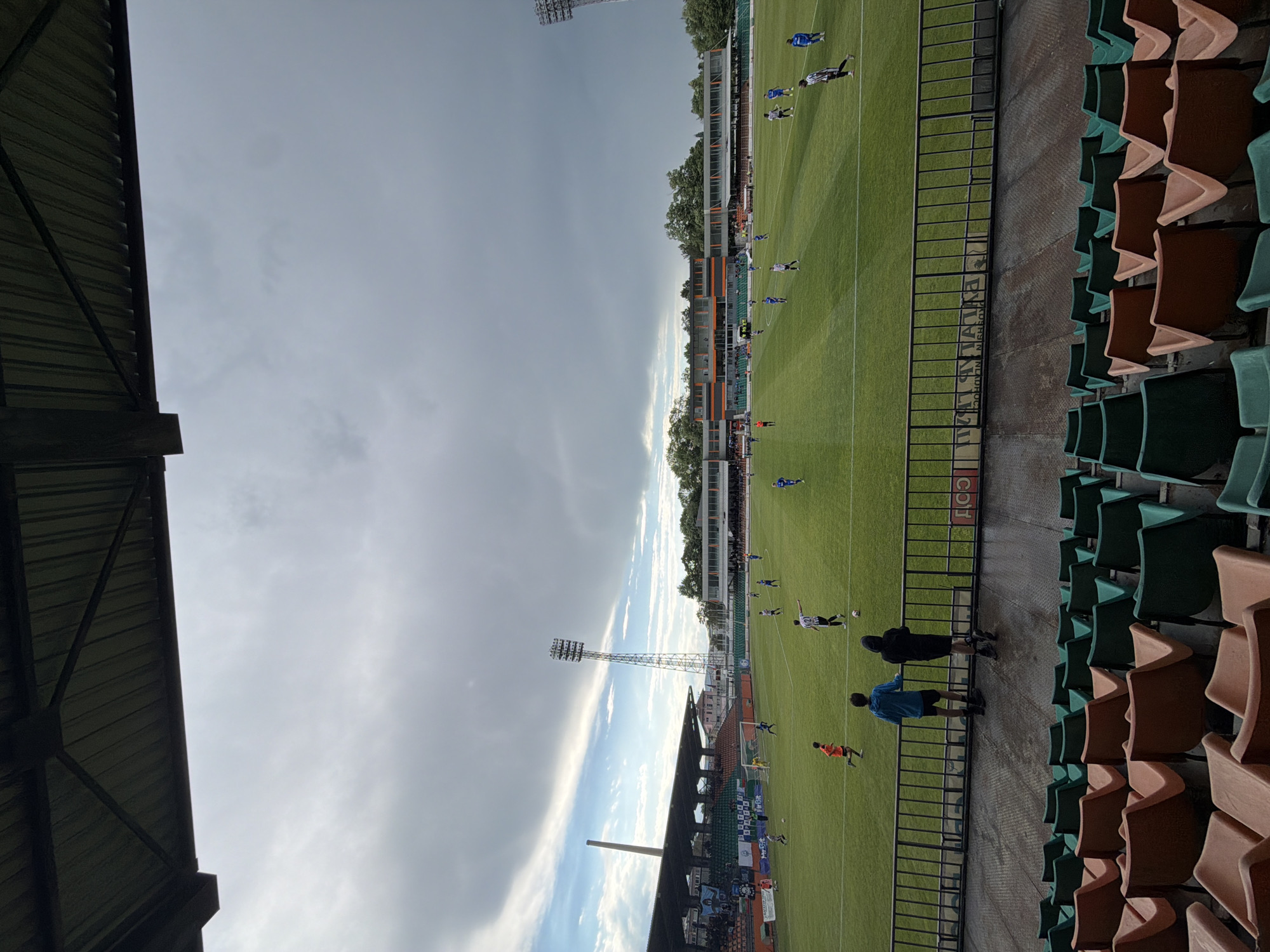 A soccer game is in progress on a green pitch, viewed from stadium seating. Spectators in the foreground watch the game, standing along the barrier between the seating and the field. The stadium seats are colored in alternating rows of green and brown. Overhead, the sky is partly cloudy. The stadium lighting towers are visible. This is most likely the Lovech Stadium, located in Lovech, Bulgaria.