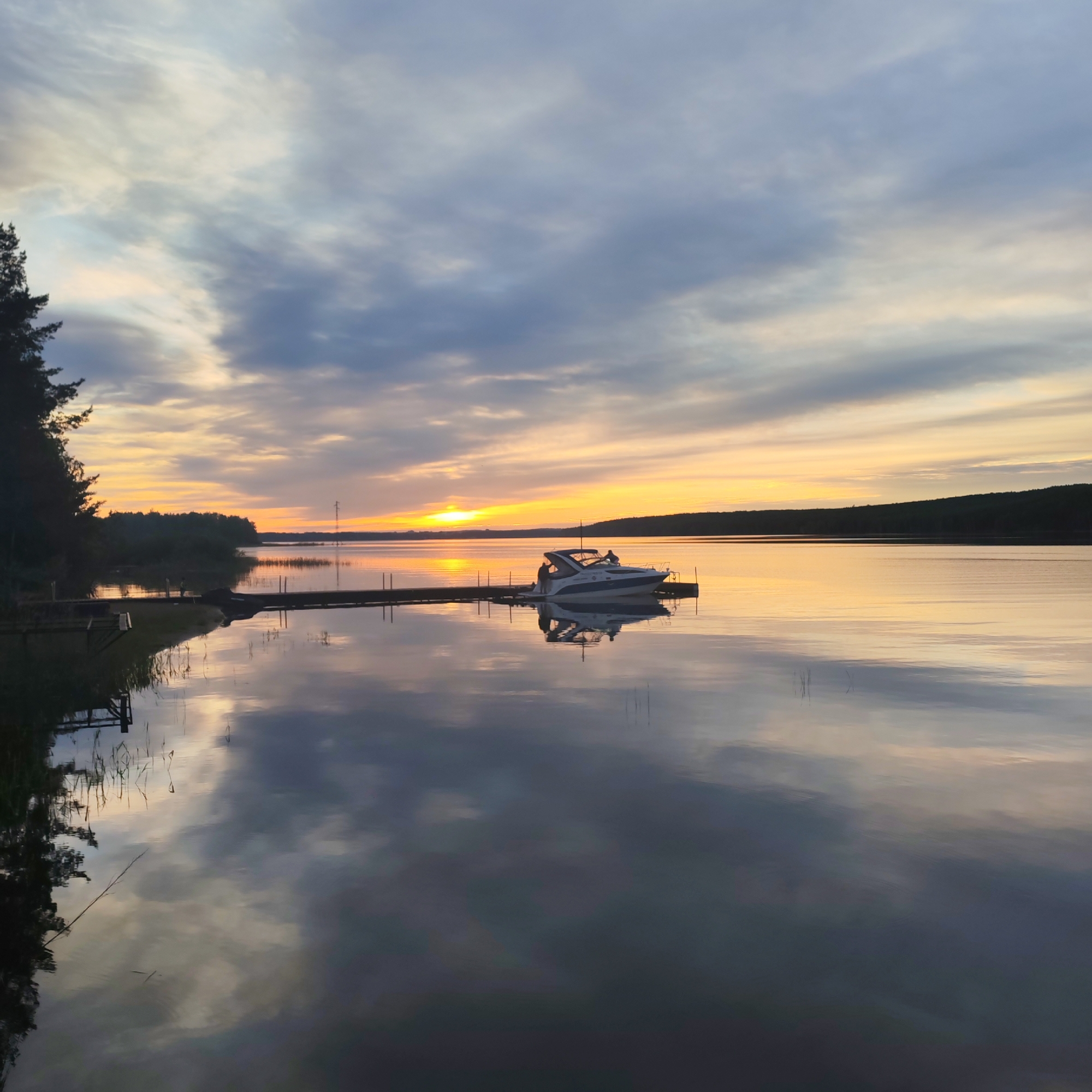 The image depicts a tranquil scene on the Kama Reservoir in Chaykovsky, Russia, captured during a sunset. A motorboat is docked at a wooden pier that stretches into the calm water. The sky is filled with a mix of gray and orange clouds, reflecting in the water. Along the shoreline, dense vegetation and trees are visible. The water is very still, creating a mirror-like effect of the sky and surrounding landscape. The lighting suggests either sunrise or sunset, with a warm glow illuminating the horizon.