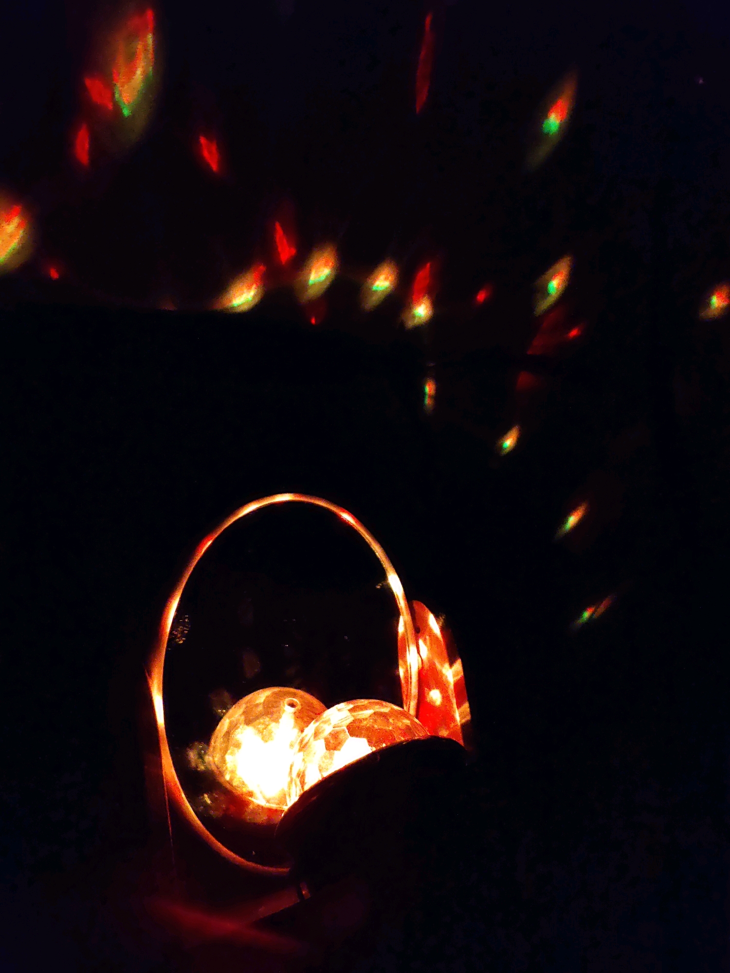 The image shows a close-up view of two illuminated disco balls in a dark environment. The disco balls are bright and appear to be made of glass with a reflective surface, possibly covered in mirrors. They are positioned within a circular frame, which is also illuminated with red and orange light. The background is dark, with numerous small, colored lights scattered across it, suggesting a light projection effect, perhaps from the disco balls themselves or another light source. The lighting is dramatic, with strong contrasts between the illuminated objects and the dark surroundings. The scene appears to be indoors, possibly a party or entertainment venue. The image does not contain any people or specific landmarks that would indicate a location in Rostov-on-Don, Russia.