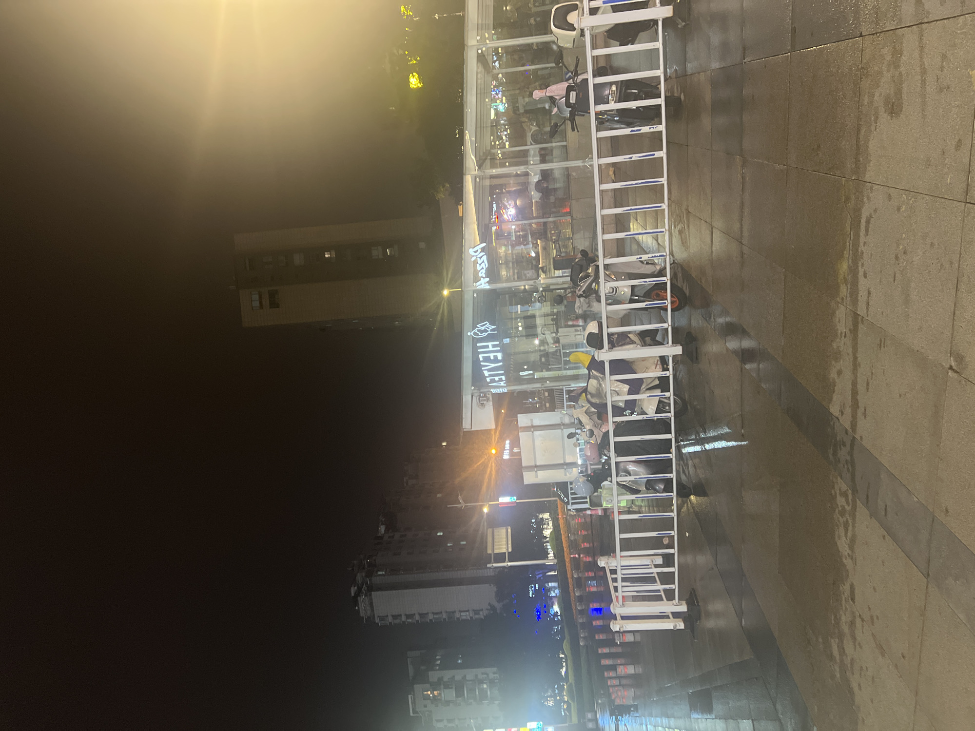 A street scene at night in Liandu District, China, shows a wet pavement reflecting lights from buildings and street lamps. In the foreground, a white metal barrier encloses several parked electric scooters. Behind the barrier, a glass-enclosed establishment with illuminated signage that appears to read "HEATYEN PIZZA" is visible, with interior lights illuminating the space. Several multi-story buildings are present in the background, some with illuminated windows. The overall atmosphere is that of a busy urban area at night, likely following a rain shower due to the wet surfaces.