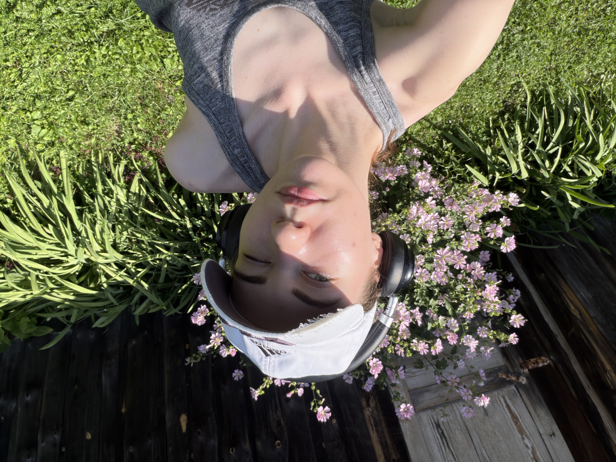 The image is a selfie taken outdoors, likely in a backyard or garden setting in Zavodoukovsk, Russia. The subject is a young woman wearing a white baseball cap and black headphones. She is looking at the camera with one eye closed in a wink. She is wearing a grey tank top. Behind her, there is a wooden fence, a bush with pink flowers, and green grass. The sun is shining brightly, casting shadows on the scene. The woman appears to be in a relaxed or playful mood.