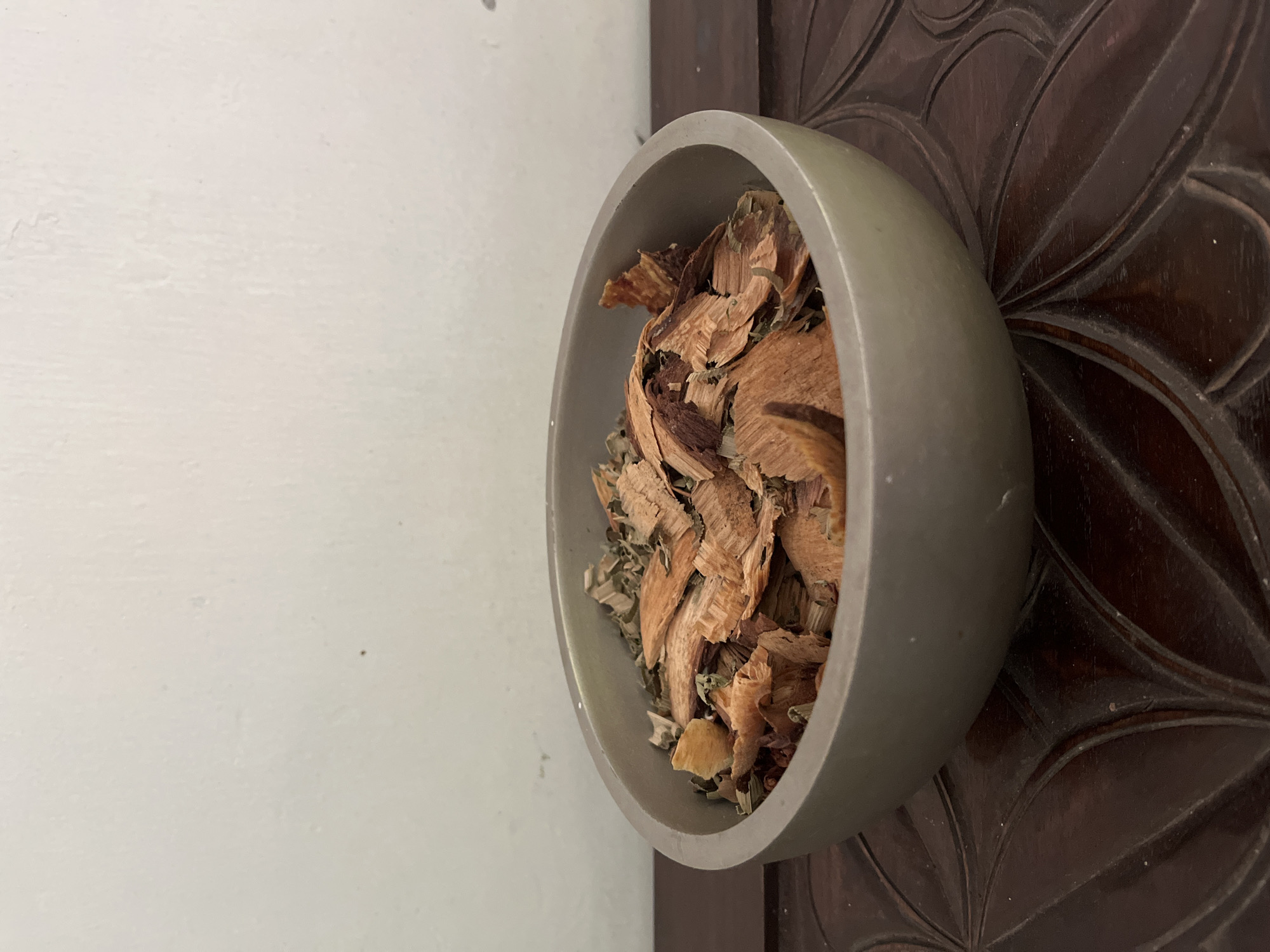 A gray bowl filled with dried bark and leaves sits on a dark wooden table with carved patterns. The background is a plain, off-white wall. The scene appears to be indoors.