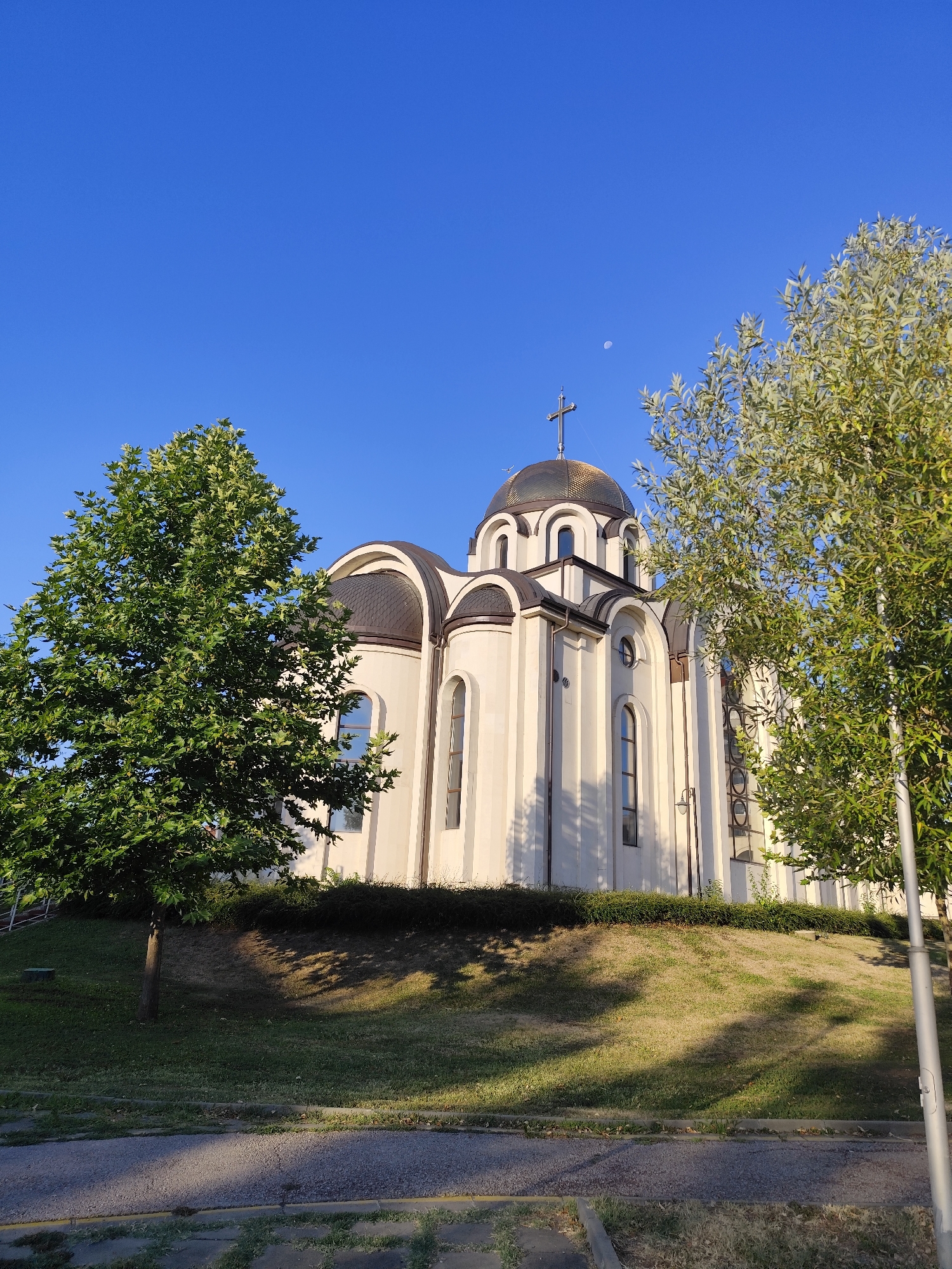 The image shows a white Orthodox church with a gold dome and cross, set against a clear blue sky. The church is situated on a grassy hill, with trees on either side of the frame. The sun is shining, casting shadows on the grass. A crescent moon is visible in the sky to the right of the church's dome. The foreground shows a paved area and some grass. This is a view of the Orthodox Church "Sveta Troitsa" located in the city of Montana, Bulgaria.