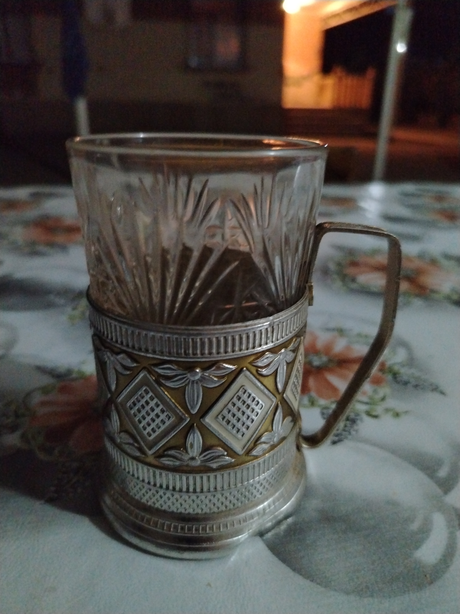A close-up, low-angle, indoor shot shows a glass cup with a metallic holder, placed on a patterned tablecloth. The cup itself is made of clear, patterned glass. The holder is ornate, featuring silver and gold-toned metalwork with intricate designs, including diamond shapes with grid patterns and floral motifs. A handle is attached to the holder. The tablecloth has a floral pattern in shades of orange and white. In the blurred background, outdoor elements are visible, including illuminated structures and dark areas, suggesting it is nighttime. The image is located in Daş Salahlı, Azerbaijan.