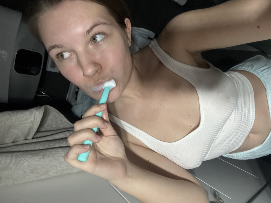 A fair-skinned woman with brown hair brushes her teeth with a turquoise toothbrush. She's wearing a white ribbed tank top and light blue shorts. The scene is dimly lit, possibly in a bathroom. Her eyes are cast to the side. A patterned blanket and a dark object are visible in the background.