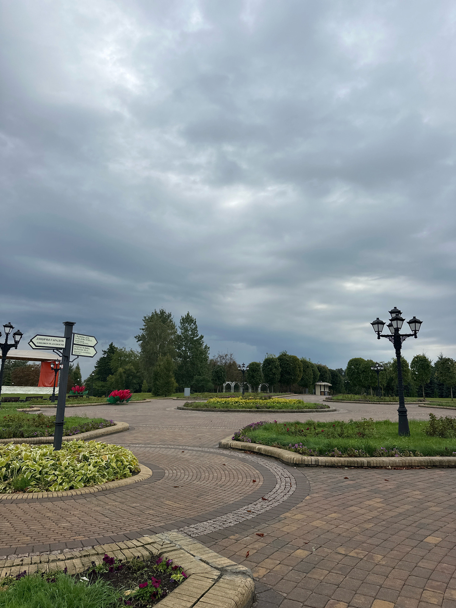 This is an outdoor, eye-level, full shot of a paved park pathway on an overcast day. The pathway is made of interlocking bricks in shades of brown and tan. In the foreground, there are several circular garden beds with green foliage and small purple flowers, bordered by yellow brick. To the left, there is a black lamppost with three lights and a signpost with directional arrows pointing left and right. One arrow reads "CHILDREN PLAYGROUND" in English and "ДЕТСКИЙ ГОРОДОК" in Russian. Further back, there is a line of trees and a raised flower bed with large red sculptural flowers. In the background, there are more trees, a small building, and white arched trellises. The sky is heavily clouded with patches of lighter gray and darker storm clouds. No people are visible in the image. The scene appears to be in a public park.