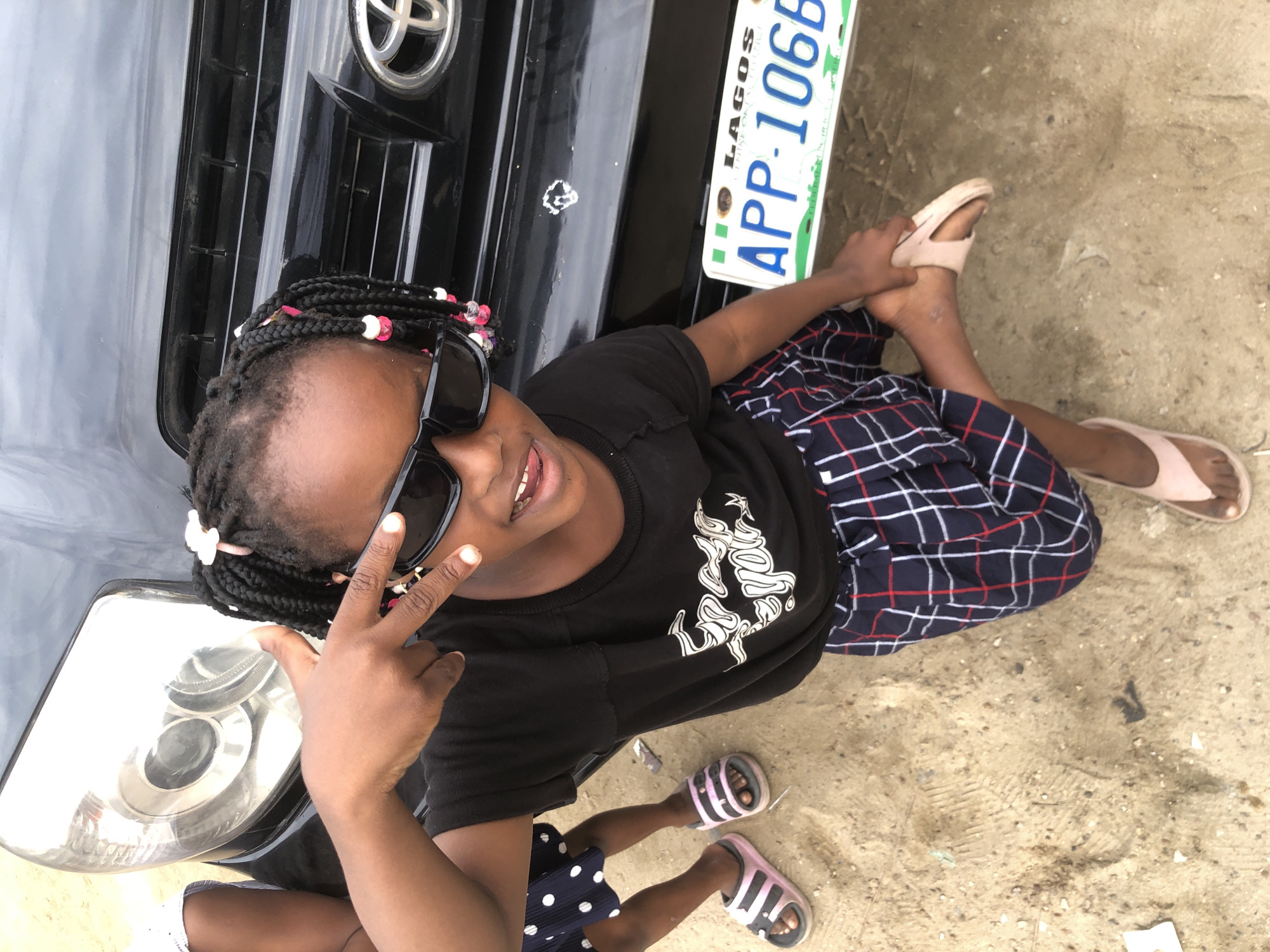 A young Black girl, appearing to be approximately 5-7 years old, stands outdoors on sandy ground near a black vehicle. She has dark skin, braided hair adorned with pink and white beads, and wears black sunglasses, a black t-shirt with white lettering, and a blue, white, and red checkered skirt. Her right hand is raised, fingers forming a peace sign, and her left foot is lifted, resting on her right ankle. She is wearing light pink sandals on both feet. Next to her, another child's legs are visible, wearing a dark blue skirt with white polka dots and pink and white striped sandals. A Nigerian license plate, "APP-106B," is visible on the rear of the vehicle. The scene is likely in Ado, Nigeria, given the license plate and the general environment.