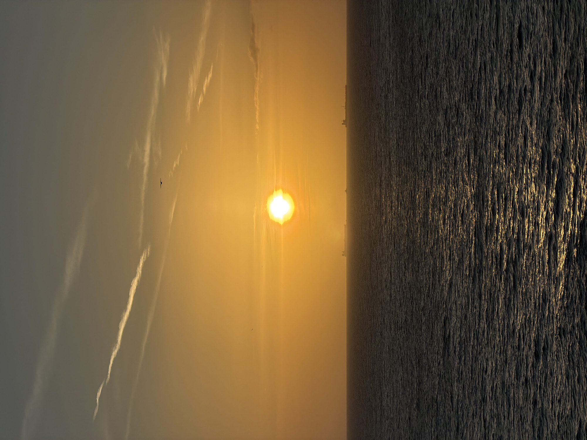 The image depicts a sunset over water. The sun is a bright, yellow orb in the center of the frame, emitting rays of light. The sky is a hazy gradient of yellow and gray, with faint contrails visible. The water is dark and textured, with reflections of the sunlight creating streaks of gold. A small bird is visible in the sky. The location is not identifiable from the image.