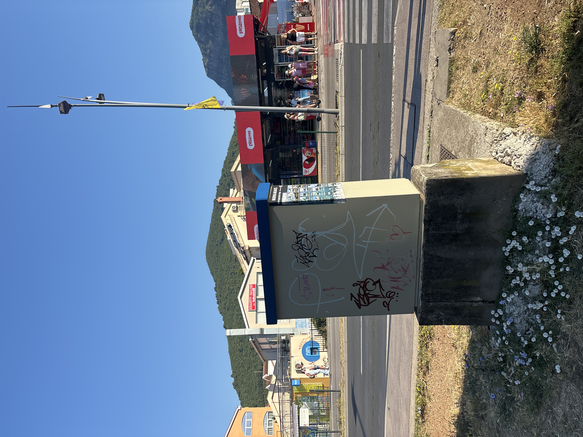 The image shows a street scene in Budva, Montenegro. In the foreground, a utility box is covered in graffiti. To the right, a pedestrian crossing leads to a row of shops with red awnings and prominent "plazma" branding. A lamppost with lights and a yellow flag stands near the shops. Several people, including women and children, are visible near the shops and on the pedestrian crossing. In the background, buildings and green hills under a clear blue sky suggest an urban environment with a mountainous backdrop. The overall scene depicts a sunny day in a public area.