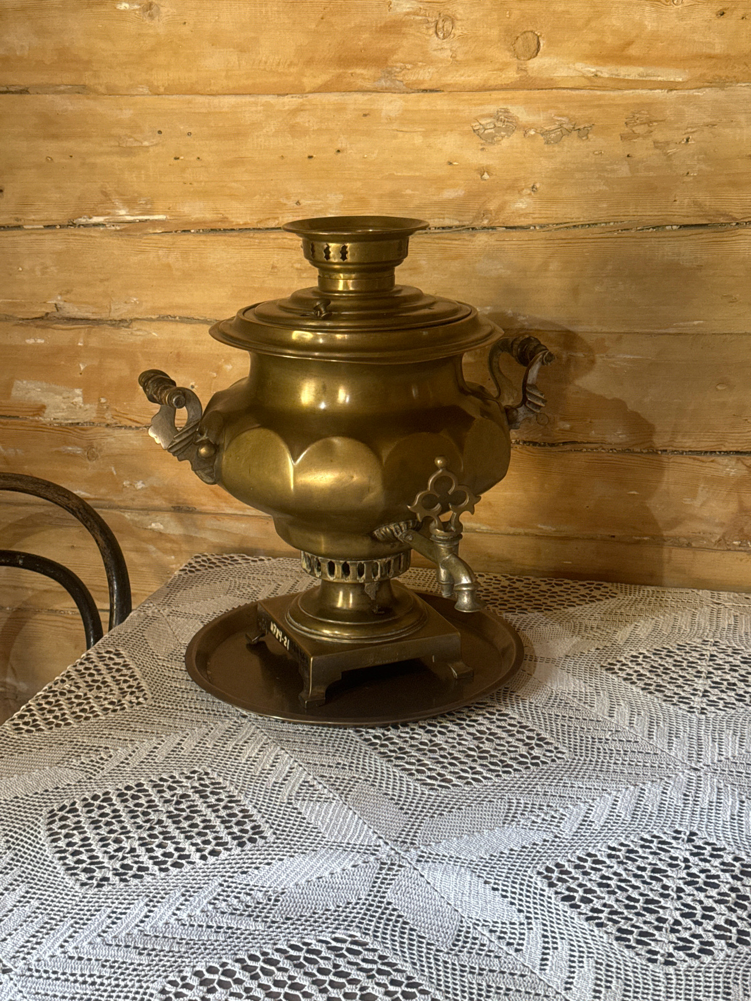 An antique brass samovar sits on a table covered with a white, lace-like tablecloth. The samovar is ornate with handles on the sides and a spigot. The background is composed of wooden planks, suggesting an indoor setting, possibly a rustic or historical establishment in the city of Taltsy, Irkutsk Oblast, Russia. A portion of a dark metal chair is visible to the left. The lighting is warm and casts shadows.