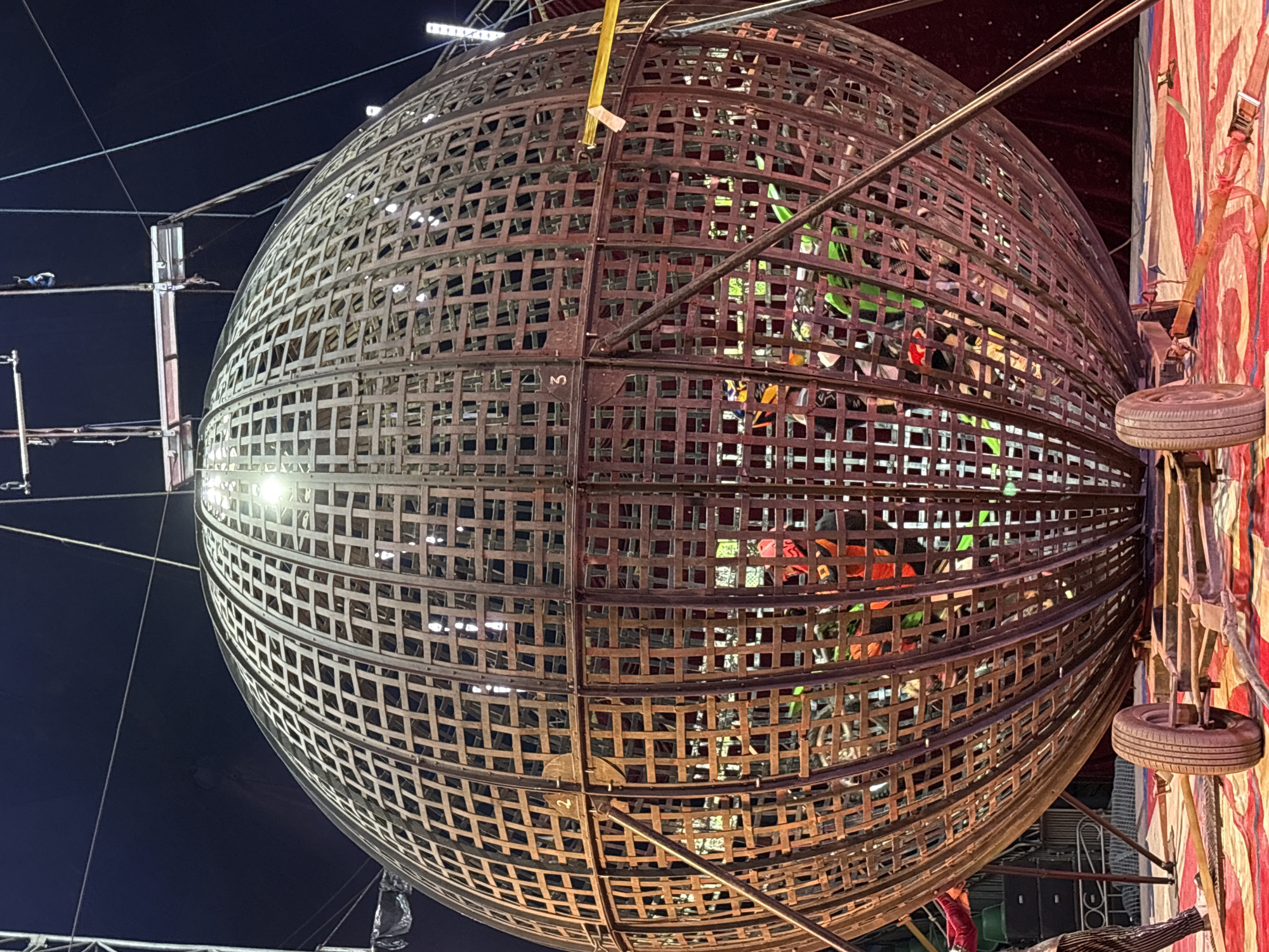 The image shows a metal globe-shaped structure, likely a "Globe of Death" performance cage, illuminated by bright spotlights. Inside the globe, there are indications of people wearing colorful clothing and possibly riding motorcycles. The globe is made of interconnected metal bars creating a grid pattern. The backdrop includes a dark night sky on one side and a red and white striped tent-like structure on the other. A tire is mounted next to the tent structure. The location is identified as Tejalpa, Mexico.
