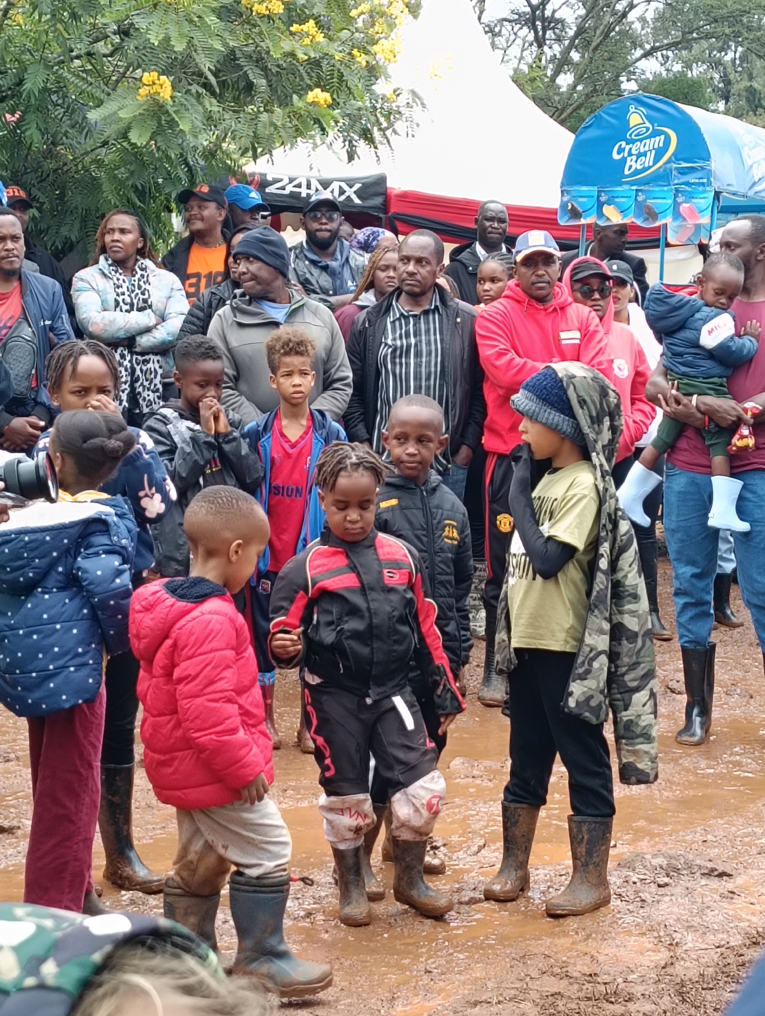 A group of people, including adults and children, are gathered in a muddy area. The adults are standing while the children are standing and interacting with each other. Some of the children are wearing rubber boots. In the background, there's a "Cream Bell" branded structure. The location is Nairobi, Kenya.