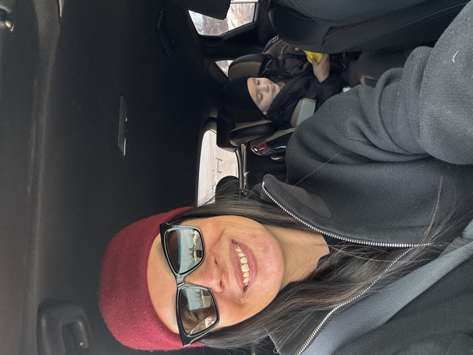 An adult female takes a selfie from the driver's seat of a vehicle in Pavlovsky, Russia. She wears a dark red beanie, black-framed sunglasses reflecting a snowy outdoor scene, and a dark zippered jacket with a grey seatbelt. Her expression is a smile, revealing teeth. In the rear passenger seat, a young child sits in a grey and black safety seat, wearing a black beanie and a dark puffy jacket. The child holds a yellow object, directing their gaze downwards. The vehicle's interior features a dark headliner. Through the rear window, a sparse, light-colored landscape consistent with winter conditions is visible, including a horizontal structure in the distance.