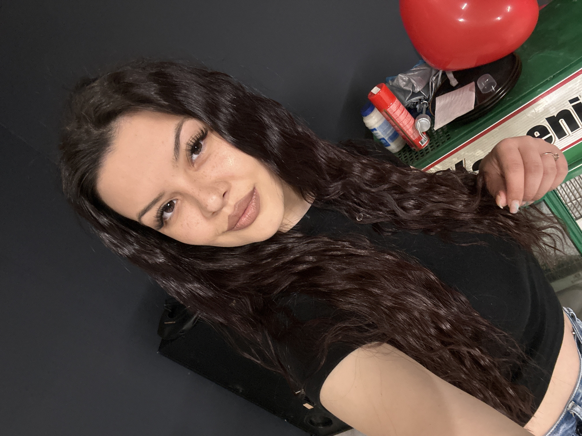 A woman with long, dark, wavy hair is shown in a selfie. She is wearing a black top and appears to be indoors. In the background, there's a green and white box, a red balloon, and some household items. The setting does not clearly indicate a specific location within Sliven, Bulgaria.