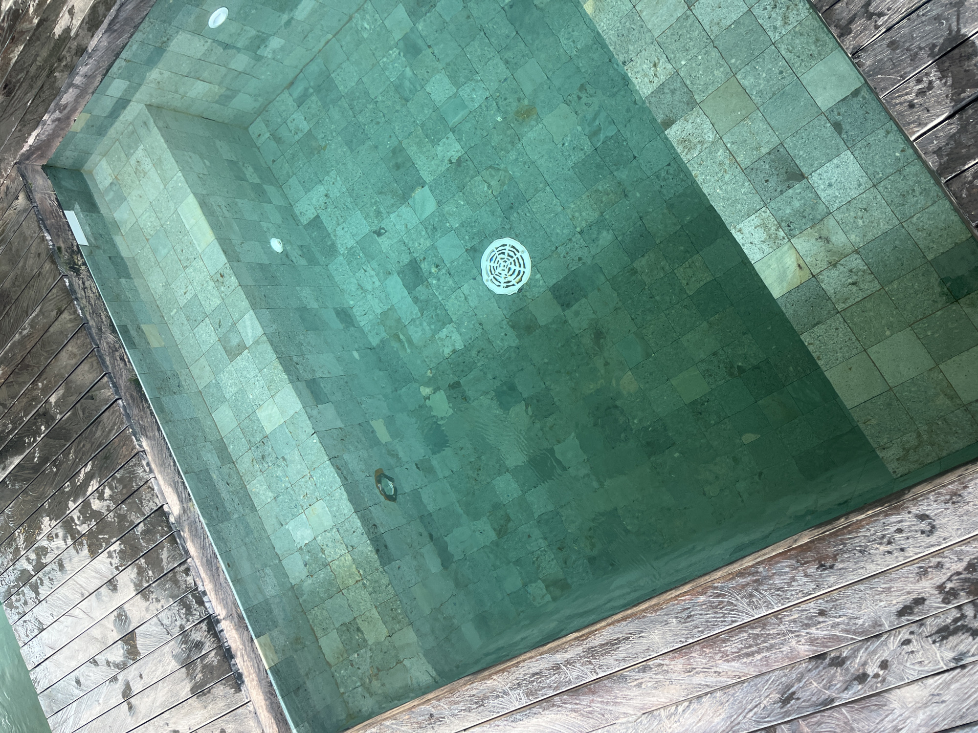 The image depicts a rectangular swimming pool filled with turquoise water. The pool bottom and walls are covered in green-toned, roughly square tiles. A circular drain is visible in the center of the pool. Two recessed lights are positioned in the upper section of the pool walls. The pool is bordered by weathered wooden decking on two sides. The image was taken in North Kuta, Indonesia.