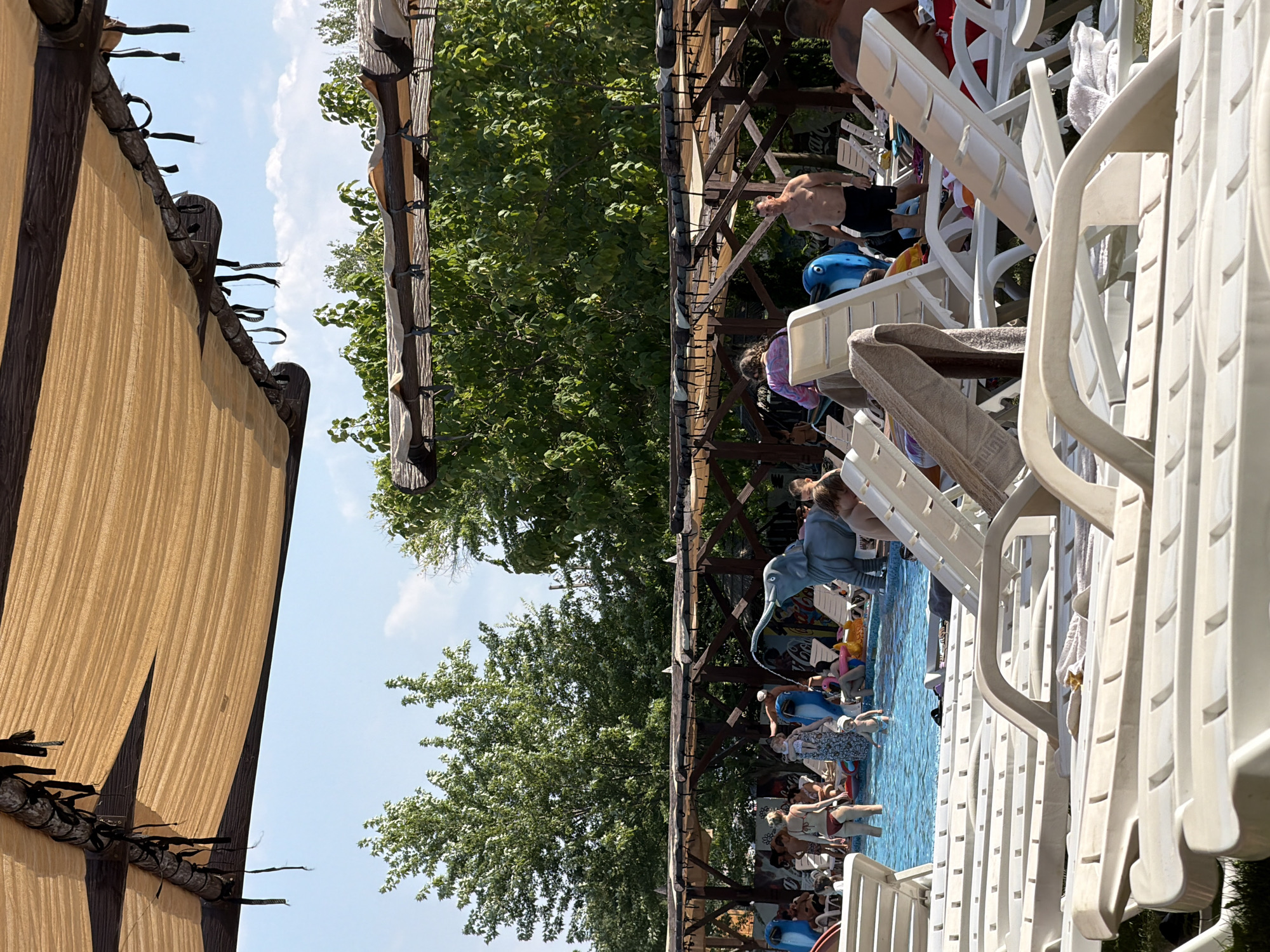 The image depicts a busy outdoor swimming pool area on a sunny day, likely at a resort or water park. In the foreground, several white plastic lounge chairs are stacked and arranged, some with towels draped over them. Beyond the chairs, a bright blue swimming pool is visible with several people enjoying the water. A large, grey inflatable elephant with a trunk spraying water is a prominent feature in the pool. Other inflatable toys, including a pink flamingo, are also present.
Numerous individuals are seen around and in the pool. Some are lounging on chairs, while others are wading or playing in the water. Children are also present, with one small child standing near the pool's edge. The background features lush green trees and a wooden canopy structure providing shade over parts of the seating area and pool. The sky is a clear blue with a few white clouds. The scene suggests a lively and recreational atmosphere. This location appears to be in Sociteni, Moldova, a village near Chisinau known for its recreational facilities, including water parks.