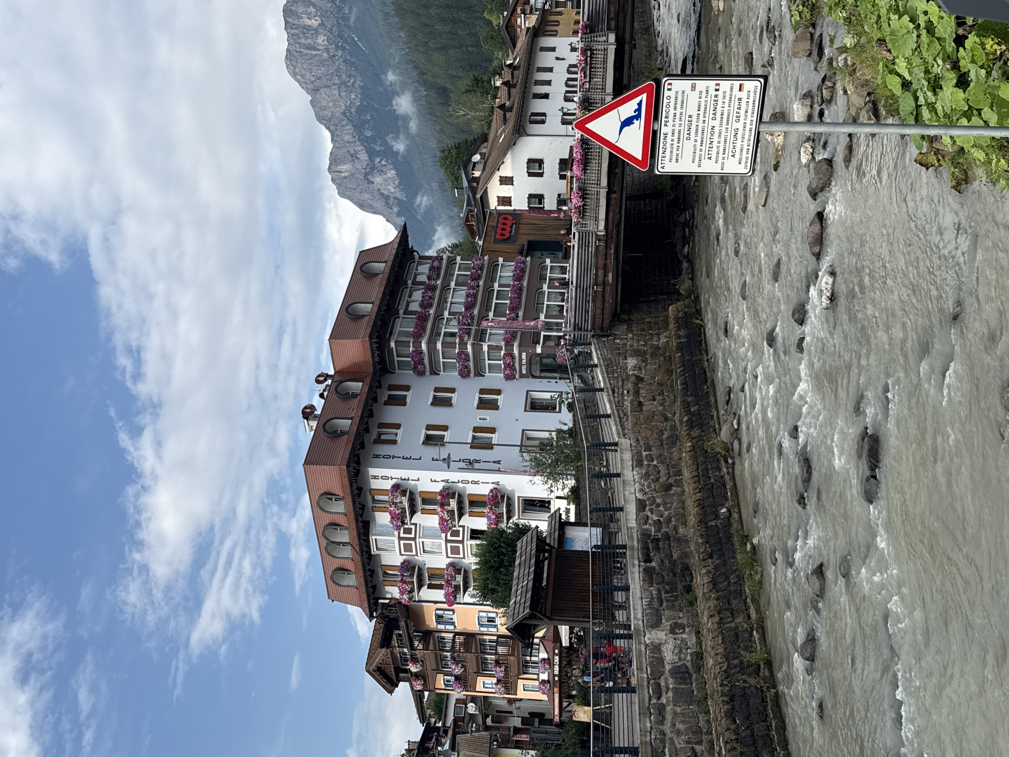 The image captures a picturesque scene in Moena, Italy, with the Fassa Creek flowing prominently in the foreground. The creek is characterized by its rocky bed and fast-moving, murky water. A stone embankment lines the creek, separating it from a walkway with a metal railing.
Behind the embankment, a pedestrian bridge spans the creek, adorned with numerous potted flowers. On the left side of the bridge, the "HOTEL FALORIA" is clearly visible, a multi-story building with a distinctive red mansard roof and dormer windows. The hotel features balconies filled with vibrant pink flowers, adding a splash of color to the facade. Next to it, another building with a similar architectural style and balconies with flowers is visible.
Further down the street, other buildings can be seen, including a "Coop" market. The background is dominated by a majestic mountain range, partially shrouded in mist, suggesting a scenic alpine location.
In the foreground, to the right of the bridge, a triangular warning sign is prominently displayed on a metal pole. The sign, featuring a red border, has an illustration of a powerful wave and text in multiple languages, including Italian and English, warning of potential dangers such as sudden flood waves.
Several people are visible on the walkway and near the buildings, appearing to be tourists or locals engaged in everyday activities. The sky above is partly cloudy, with patches of blue sky visible between the clouds. The overall atmosphere is one of a charming mountain town in Italy, with a blend of natural beauty and urban architecture.
