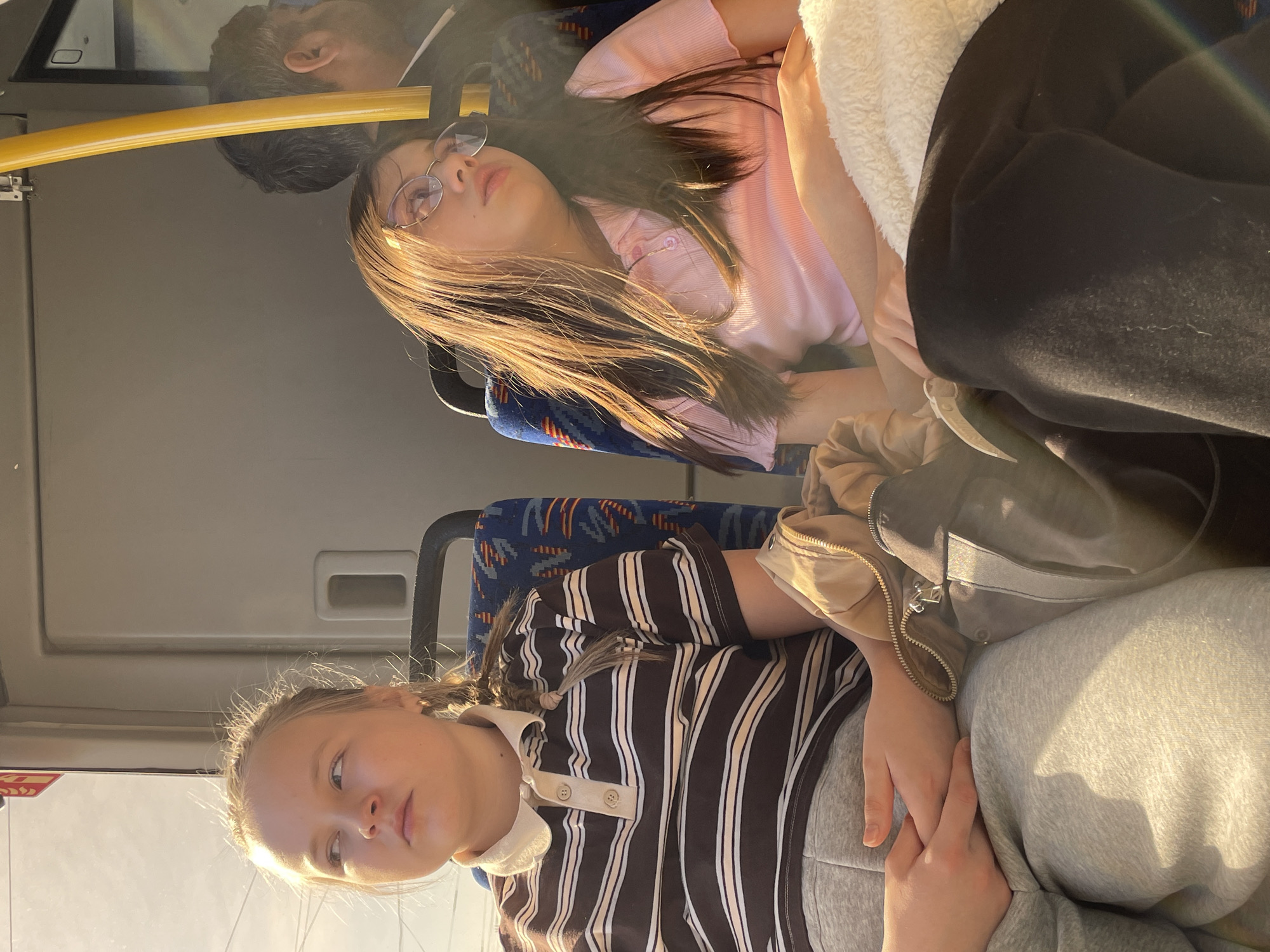 Two young females are seated inside a public transportation vehicle, possibly a bus. One female is in the foreground, wearing a brown and white striped shirt and gray sweatpants. She has light skin and her blonde hair is in pigtails. Her hands are clasped together in her lap.
The other female, located in the background, has light skin and long brown hair. She is wearing a light pink polo shirt and glasses. She is sleeping or resting with her eyes closed. The interior of the bus is visible, including the blue patterned seats, a yellow handrail, and window frames. An older male is above the female with long brown hair. The location is unknown.