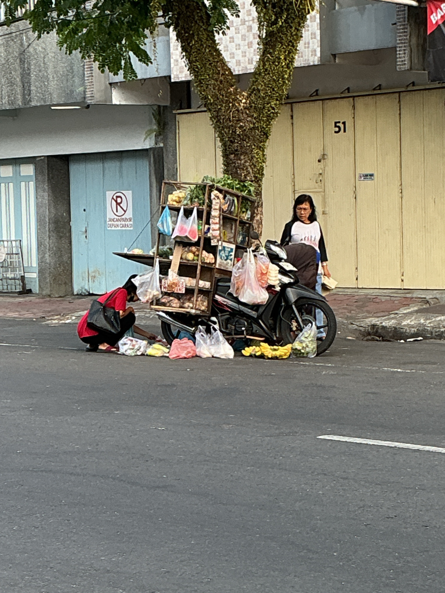 In a street scene in Magelang, Indonesia, a woman in a red shirt is crouched down next to a motorcycle laden with a cart filled with produce and other items for sale. She is arranging items on the ground. A second woman in a white and black shirt stands behind the motorcycle, looking towards the camera. The motorcycle is parked on the side of a street with buildings in the background, one of which has the number "51" on it. A tree with dense foliage is positioned between the two women and the building. The ground is paved with asphalt, and a sidewalk is visible behind the motorcycle. The scene appears to be a street vendor preparing to sell goods.