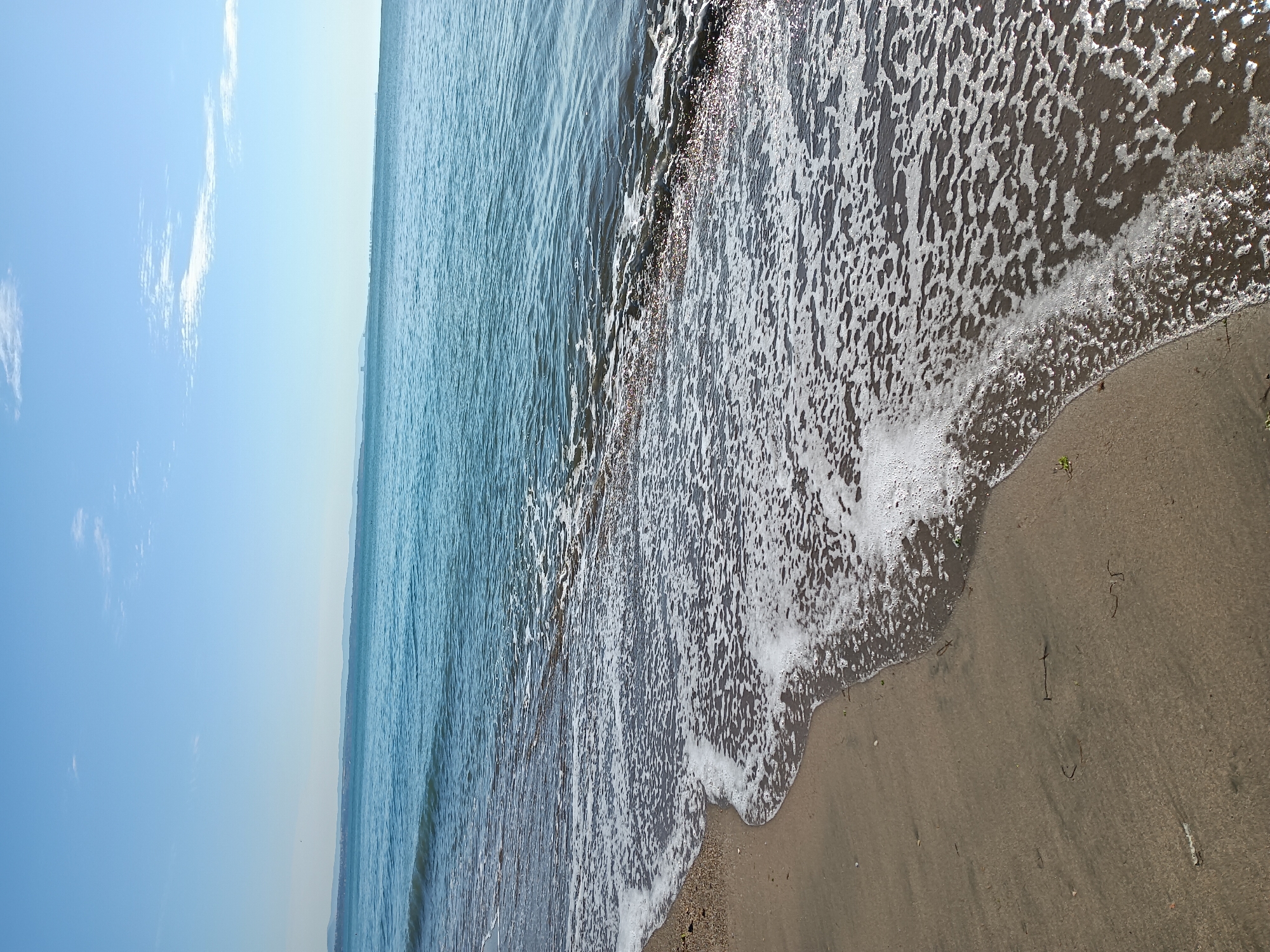 The image depicts a beach scene with the Black Sea. The camera is positioned close to the water's edge, looking out towards the horizon. The water is calm with gentle ripples, and a small wave is washing ashore, creating white foam on the wet sand. The sand is a light brown color, and a few pieces of dark seaweed are scattered on it. In the distance, a hazy outline of land can be seen along the horizon, suggesting a coastline. The sky is a clear, light blue with a few wispy white clouds. The sun appears to be high in the sky, casting a bright light that creates reflections on the water and makes the foam sparkle. The overall mood is peaceful and serene. The location is implied to be Burgas, Bulgaria, a city known for its beaches on the Black Sea coast. There is no visible text in the image.