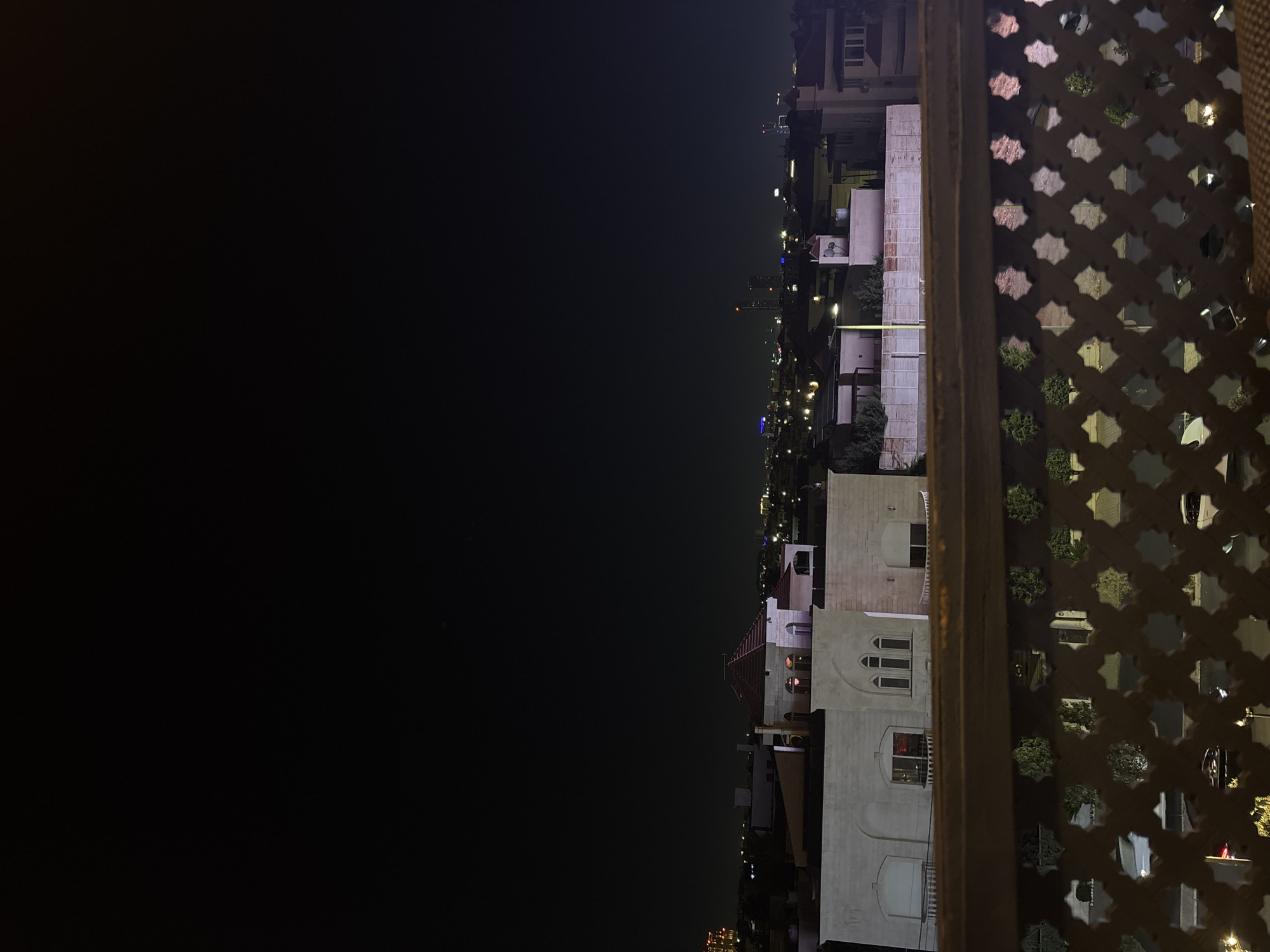 The image is a nighttime view of a cityscape, likely Amman, Jordan, taken from an elevated position. In the foreground, a decorative railing with a starburst pattern is visible, with the tops of potted plants peeking through some of the openings. Beyond the railing, several buildings are illuminated. The architecture suggests a residential or mixed-use area. One prominent building in the mid-ground has a light-colored facade, a red-tiled roof with dormer windows, and arched windows on its upper floor. In the background, a sprawling city lights up the horizon, with taller buildings and distant lights indicating a more urbanized area. The sky is dark, suggesting it is well after sunset. No people are discernible in the image.