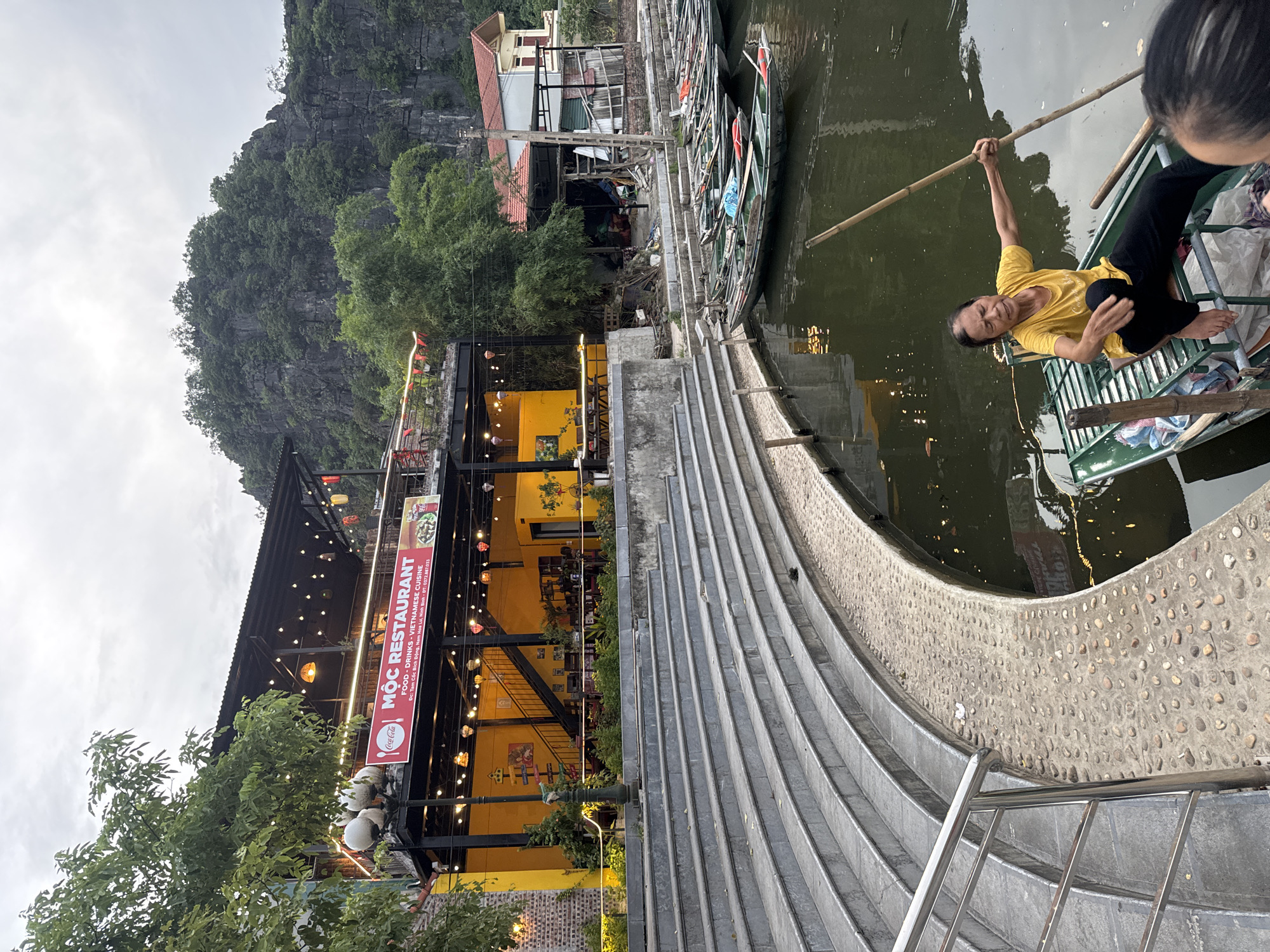 The image depicts a waterside scene in Nam Hoa Lư Ward, Vietnam, dominated by a vibrant yellow building identified as "MỘC RESTAURANT" which offers food, drinks, and Vietnamese cuisine. The restaurant features multiple levels, with the upper floor illuminated by string lights and decorative lanterns. Below the restaurant, a set of wide, curved concrete stairs leads down to the water's edge. The water is populated by numerous green, long, narrow boats docked alongside a stone embankment. In the foreground, a woman wearing a yellow t-shirt and black pants is seated in one of these boats, holding a long wooden pole, seemingly in the process of navigating or preparing to navigate the water. Behind the boats and restaurant, a lush, tree-covered rocky cliff face rises, characteristic of the karst topography of the region. The sky is overcast, suggesting a diffused light conditions.