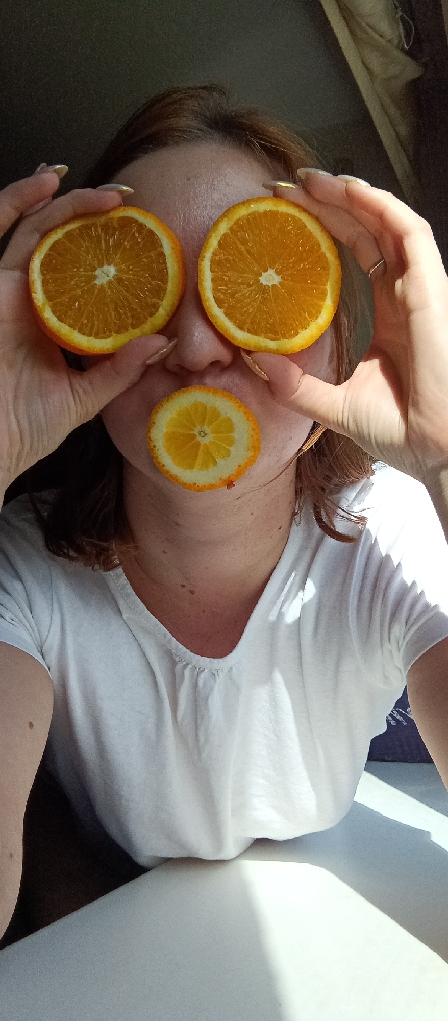 A woman is holding orange slices up to her face, covering her eyes and mouth. The woman is wearing a white t-shirt. She has light skin, with freckles on her neck. Her hair is reddish-brown. The background is indistinct, but there appears to be a window and some furniture. The location is unknown.