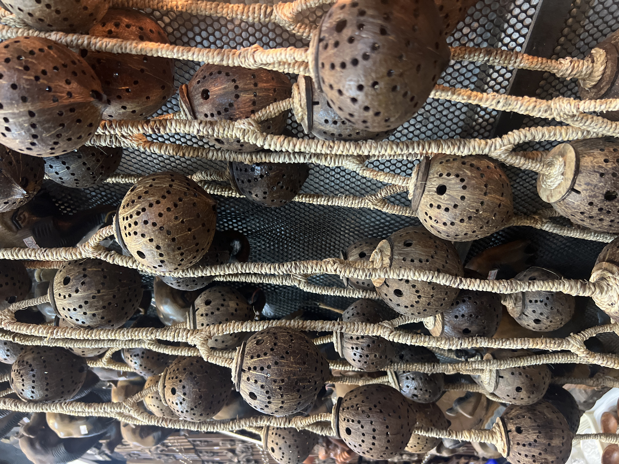 The image depicts a close-up view of numerous hanging decorative items made from hollowed-out coconuts, each perforated with small holes. These coconut ornaments are suspended by thick, braided ropes. The background features a dark, mesh-like surface, possibly a net or screen, and hints of other items, suggesting a market or shop setting. The lighting is warm and somewhat dim, casting shadows and highlighting the textures of the coconuts and ropes. This scene is characteristic of souvenir stalls found in Karon, Thailand, where such handcrafted items are commonly sold. There are no people or animals visible in the frame.