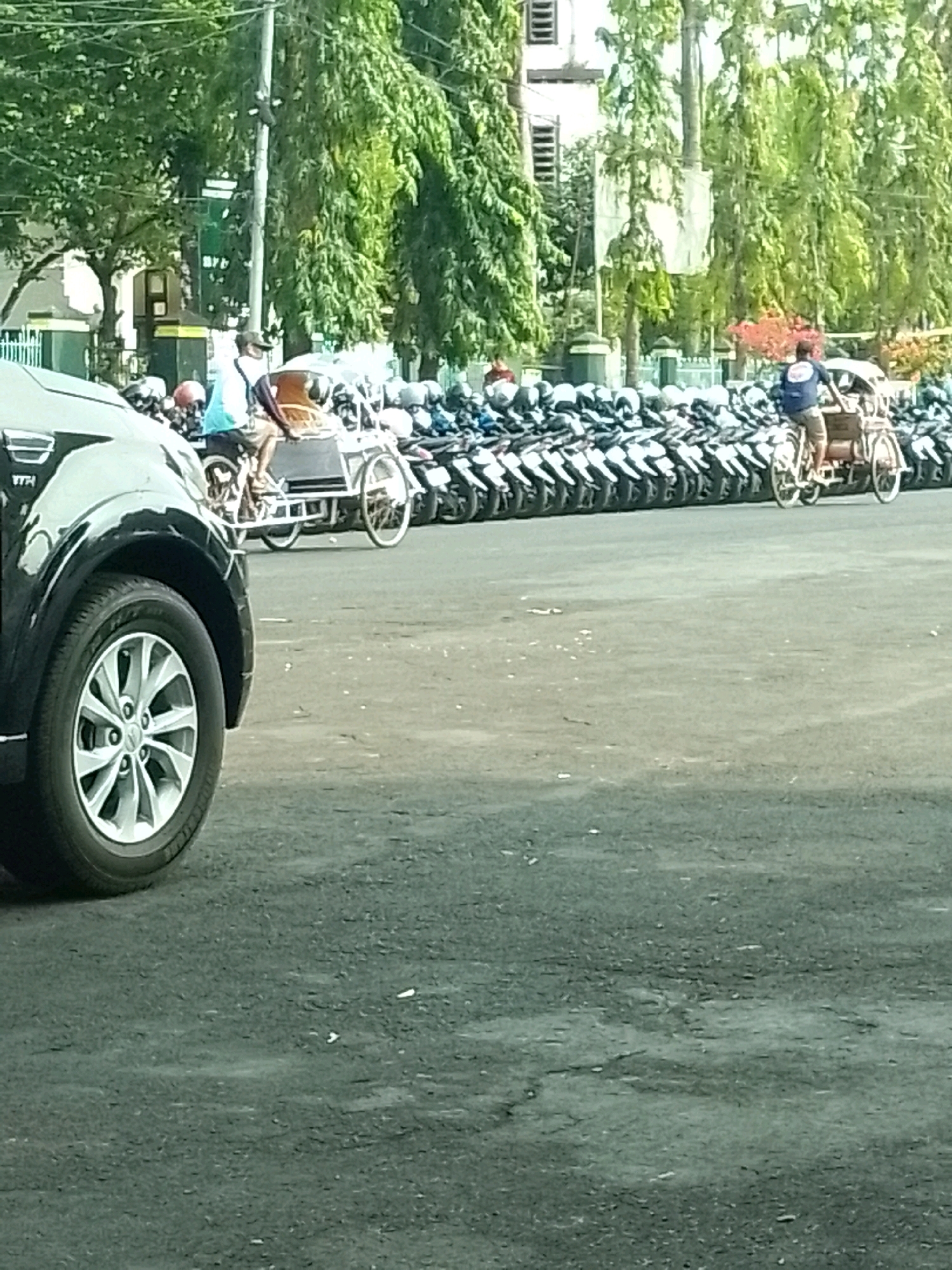 The image shows a street scene in Jember, Indonesia. In the foreground, the front passenger side of a black SUV is visible. Behind the SUV, a person is riding a pedicab, with a long line of parked motorcycles extending into the background. Trees and buildings are visible behind the motorcycles. The scene appears to be a sunny day, with good visibility. The area is paved with asphalt, and there is some debris on the ground. The overall impression is of a typical street in a Southeast Asian city, with a mix of vehicles and pedestrians.