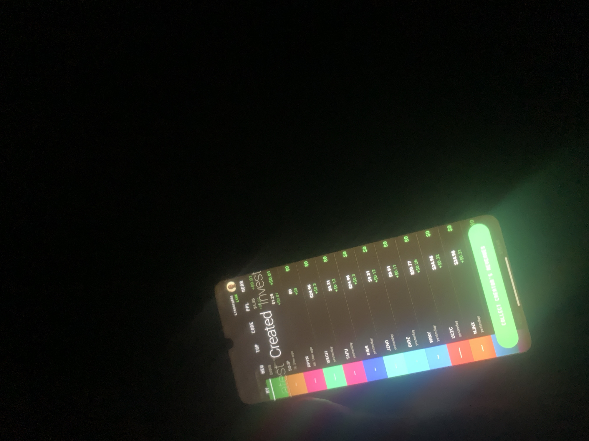 The image shows a person holding a smartphone in a dark environment. The phone screen displays a financial application with a list of investments and their corresponding values and changes. The application's name appears to be "Created Invest". At the bottom of the screen, there is a prominent green button that says "COLLECT CREATOR'S REVENUES". The background is dark, suggesting it is nighttime or an indoor setting with low lighting. There is no indication of the city of Jalingo, Nigeria in the image.