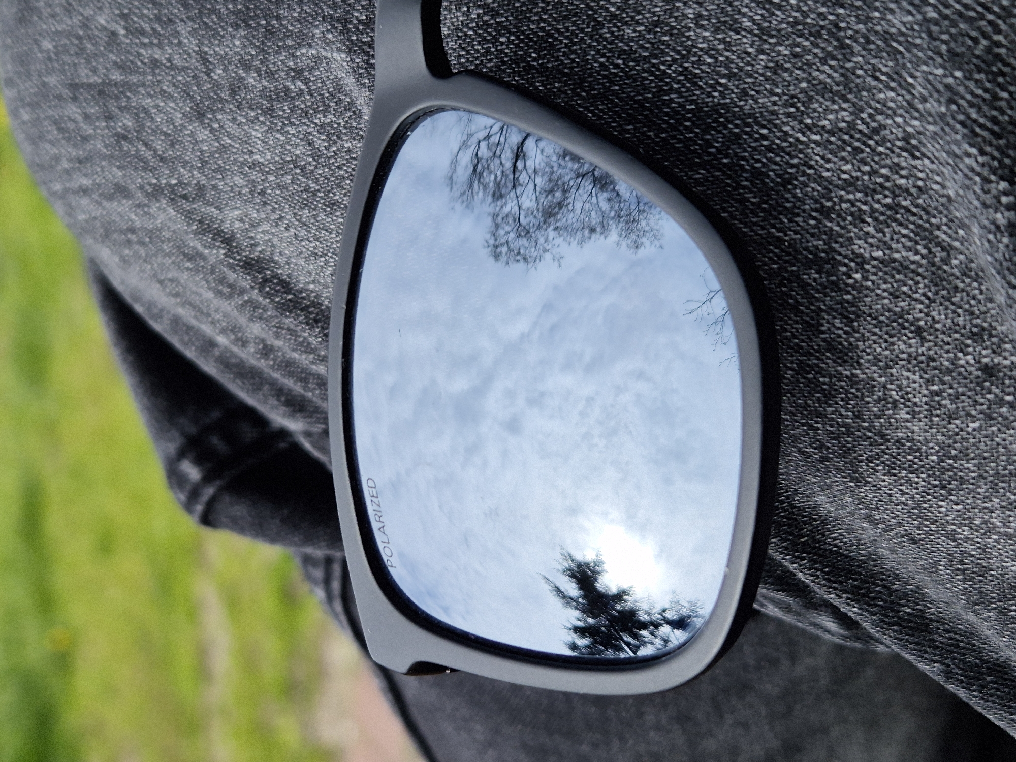 A close-up shot features a pair of sunglasses with a black frame resting against a dark gray denim fabric. The lens of the sunglasses reflects a sky with clouds and tree branches. The word "POLARIZED" is visible on the frame. Green grass is partially visible in the background on the left. The location cannot be determined to be in Varna, Bulgaria, based on the visible elements.