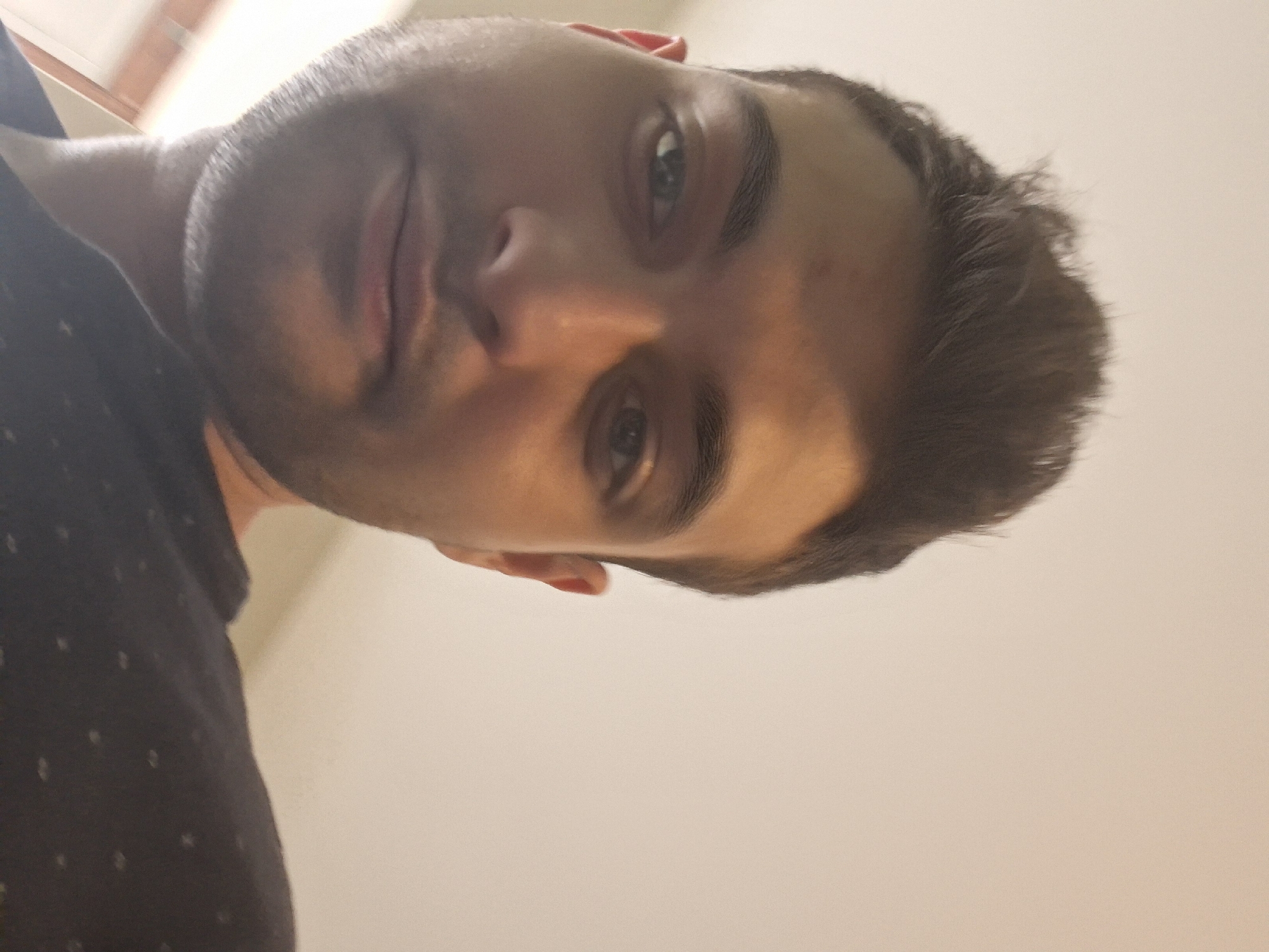 A close-up, low-angle selfie shows the face of a young man against a plain, light-colored background. He has dark, styled hair, dark eyebrows, and brown eyes. His head is tilted slightly upwards and to the left. He is wearing a dark shirt with a subtle pattern. The lighting appears to be from above and to the right, casting a slight shadow on the left side of his face. The background suggests an indoor setting, possibly a room with neutral walls and a ceiling. The image is slightly blurry, particularly in the extremities. There is no discernible context suggesting a specific location in Sofia, Bulgaria, or any indication of interaction with other individuals or objects beyond what is visible on the person.