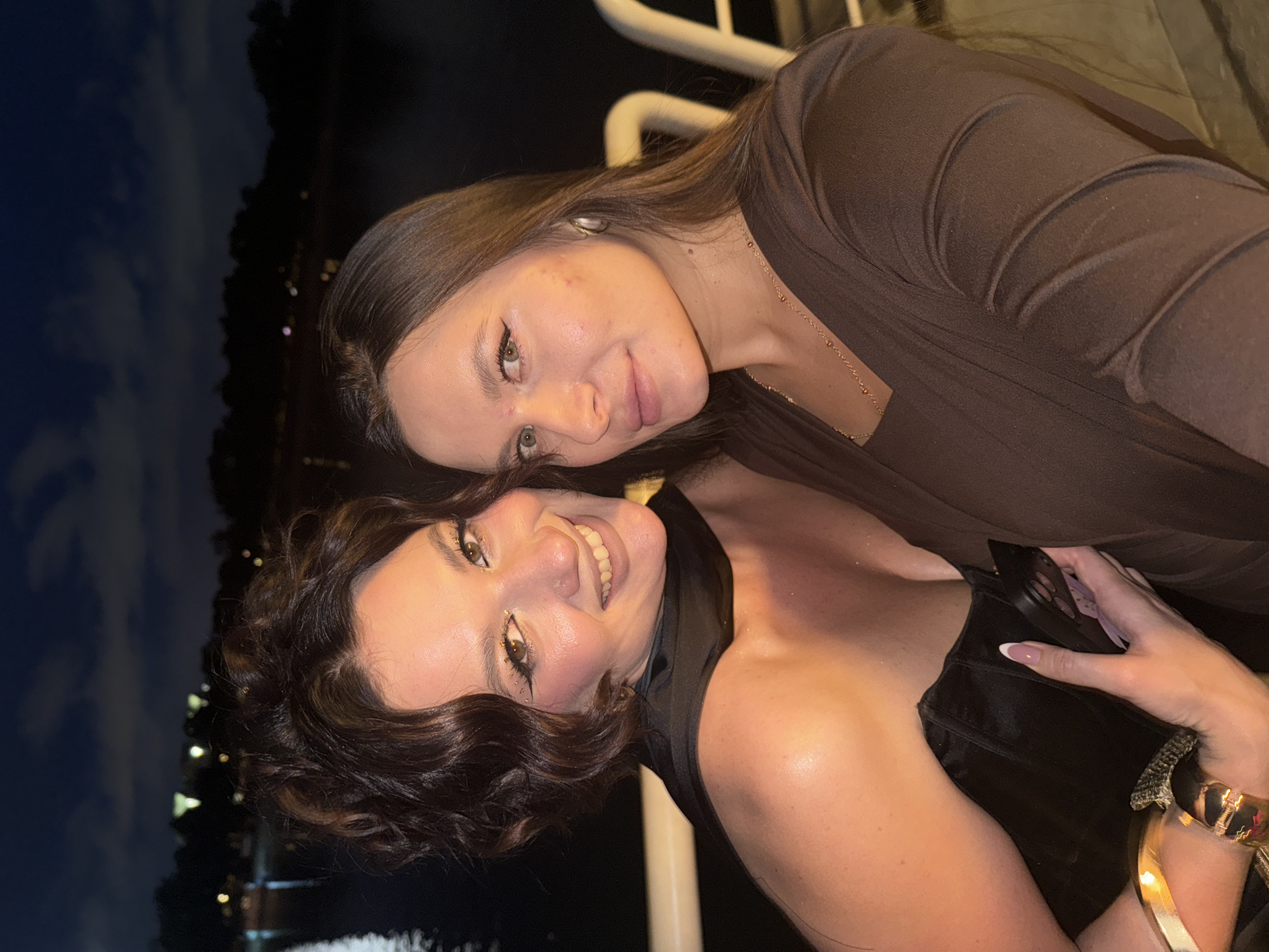 Two women are posing closely together in an outdoor setting at night, likely on a boat or near water. The woman in the foreground is wearing a black top, has dark curly hair with golden highlights, and noticeable decorative makeup around her eyes. She is smiling. The woman behind her has straight brown hair and is wearing a brown top. The background shows a city skyline with lights, suggesting a possible location like the Belgrade waterfront in Serbia, known for its nightlife along the Danube and Sava rivers. The lighting is dim, creating a night-time atmosphere.