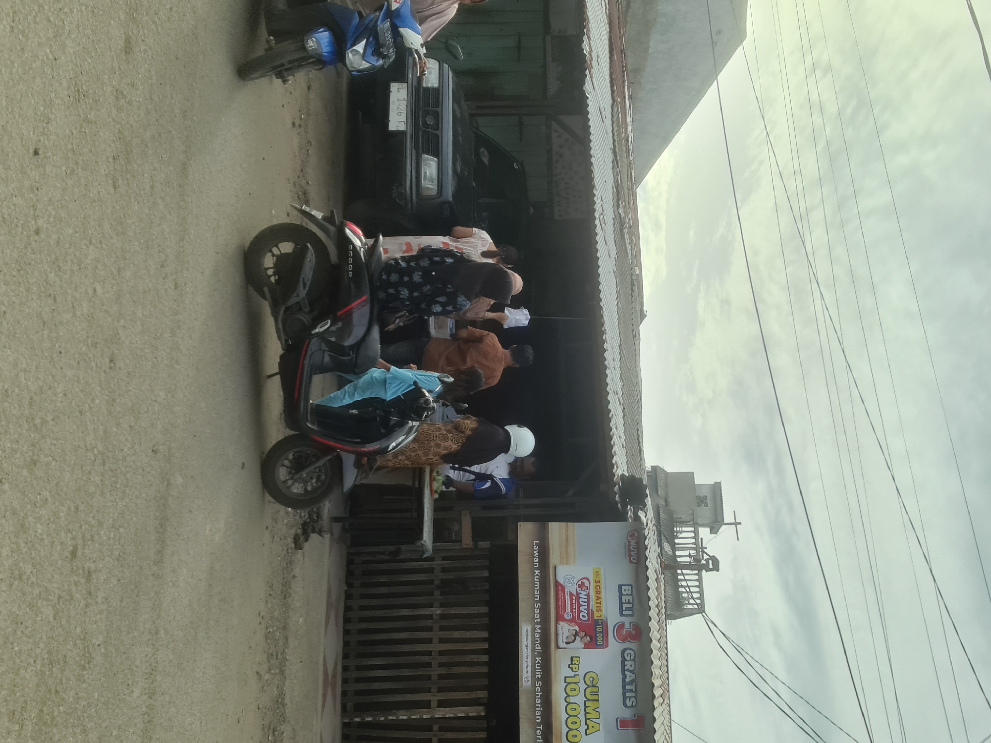 The image depicts a street scene in Lhokseumawe, Indonesia, under an overcast sky. In the foreground, two motorcycles are parked on a dirt and asphalt road. A dark-colored SUV is parked behind a makeshift stall, which has a corrugated metal roof and wooden siding.
Several people are gathered around the stall. A woman in a black hijab and patterned dress stands near the front of the stall, facing away from the camera. To her left, a child in a light blue outfit is seated on the back of a dark scooter. Behind the child, a woman in a white and orange patterned dress and a headscarf is interacting with someone at the stall. A man in an orange shirt stands to the right of the stall, facing the woman in the patterned dress. Another person, wearing a white helmet and blue and white shirt, is also visible behind the stall.
A large banner advertising "NUVO BELI 3 GRATIS 1" (NUVO Buy 3 Get 1 Free) with a promotion for 10,000 IDR is displayed on the right side of the stall. Text below the banner reads "Lawan Kuman Saat Mandi, Kulit Sehari-hari Terli..." (Fight Germs While Bathing, Daily Skin...). The environment suggests a commercial area, possibly a small shop or roadside vendor. Overhead, a network of power lines crisscrosses the sky.