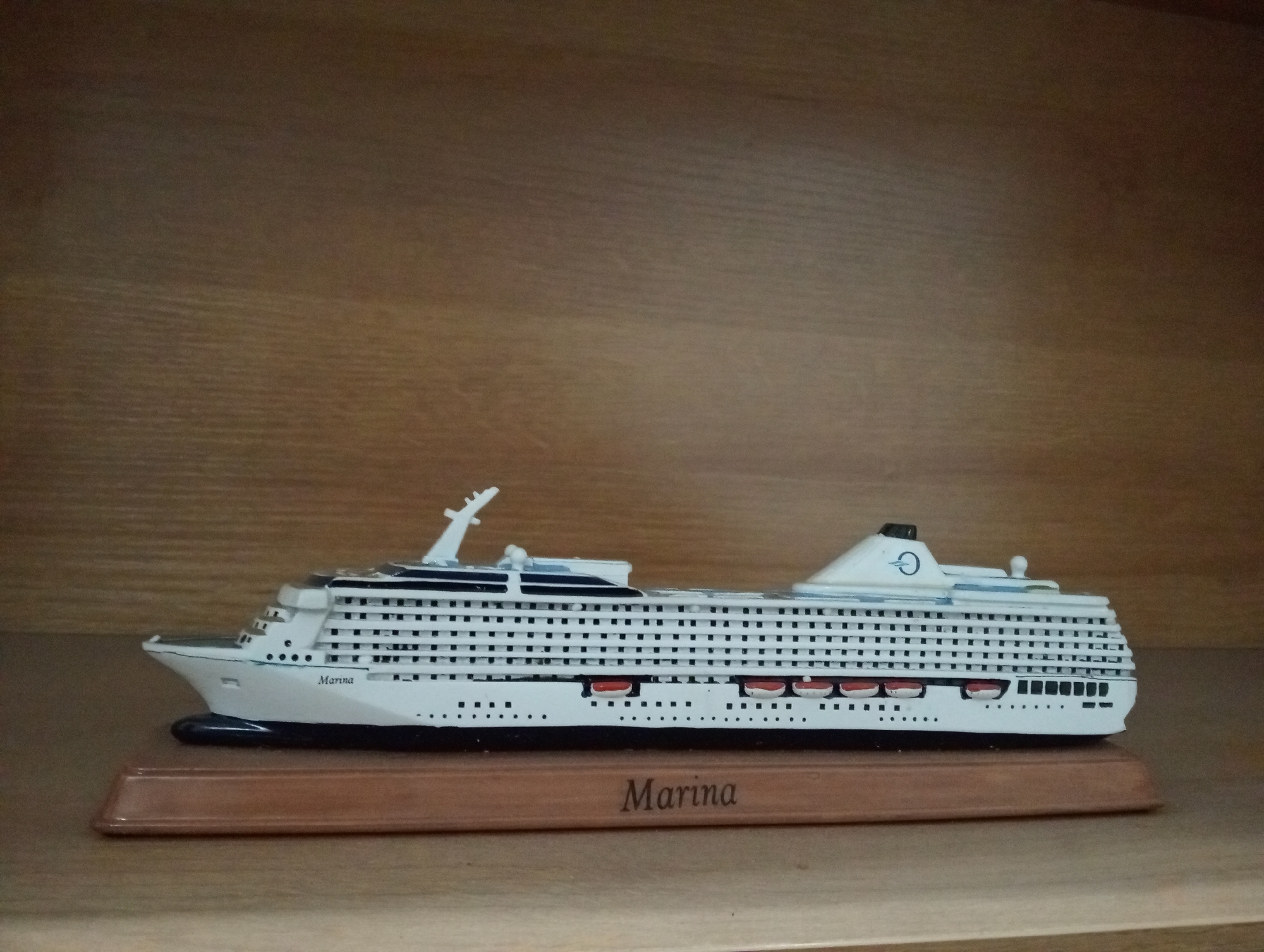 A detailed, forensic, matter-of-fact description of the image:
The image depicts a model cruise ship, labeled "Marina," resting on a wooden display stand. The cruise ship is white with black accents and features multiple decks with rows of windows. Lifeboats are visible along the side of the ship. The display stand is a light brown, elongated rectangle. The model is positioned on a wooden shelf. The background is a textured wooden surface, likely part of a bookshelf or wall paneling. There are no people or interactions visible in the image. The lighting suggests an indoor environment, possibly a display case or shelf.
The provided image does not contain any information that allows for the identification of a location in the city of Denpasar, Indonesia.