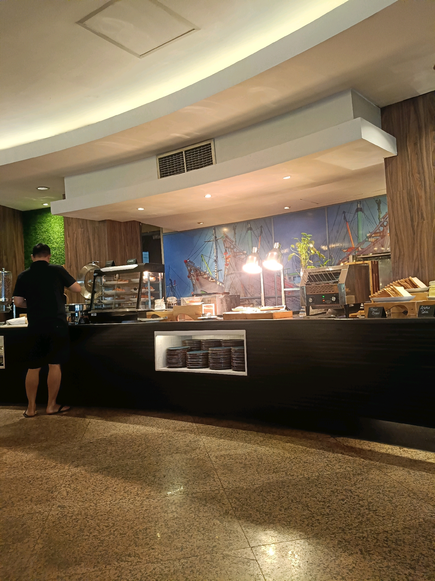 The image depicts a breakfast buffet area at a hotel in North Jakarta, Indonesia. In the foreground, a man wearing a black t-shirt and shorts stands facing away from the camera, appearing to be selecting food from a buffet station. The buffet counter is black and displays various food items, including pastries in a glass display case and a toaster with bread. Behind the counter, a mural with a nautical theme is visible. The floor is tiled with a speckled pattern. The lighting suggests it is either early morning or evening.