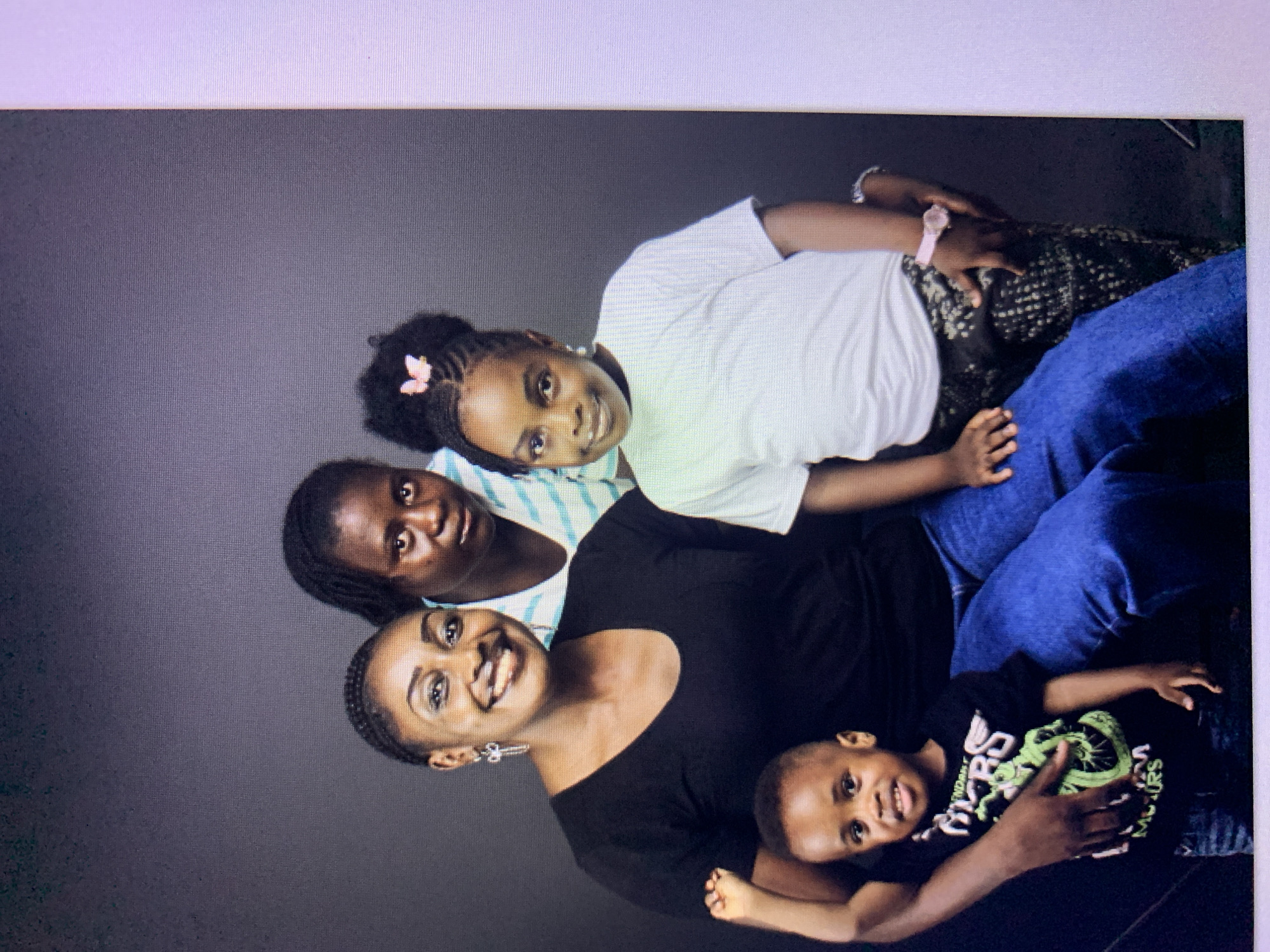 This is a studio portrait of a family of four. A woman with braided hair and a black shirt is in the front left, holding a young boy in a black t-shirt with a green graphic. Behind them, slightly to the left, is another woman wearing a striped shirt. To the right of the boy, and in front of the woman in the striped shirt, is a young girl with her hair in a bun, wearing a white t-shirt and patterned pants. The background is a plain dark gray. The image is framed in a way that suggests it was taken indoors, likely in a professional photography studio. There is no information to suggest the location is Gura Top, Nigeria.