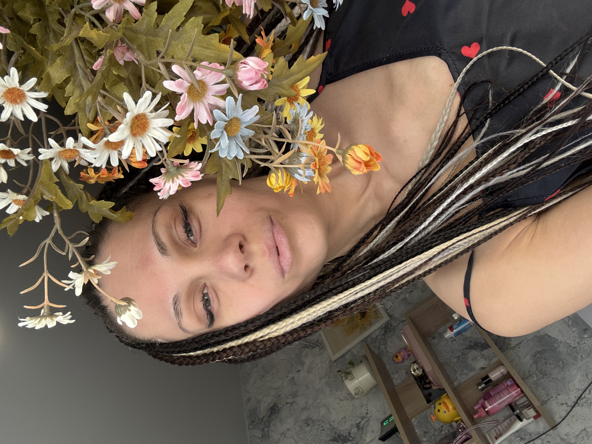 A woman lies on her side, facing the camera, with an array of artificial flowers positioned around her head. She has braided hair with blonde highlights. She is wearing a black garment with red hearts. In the background, a shelf unit is visible, containing various items, including toiletries and small trinkets. The setting appears to be indoors, possibly a room in a home in Zhukovo, Russia, as per the prompt.