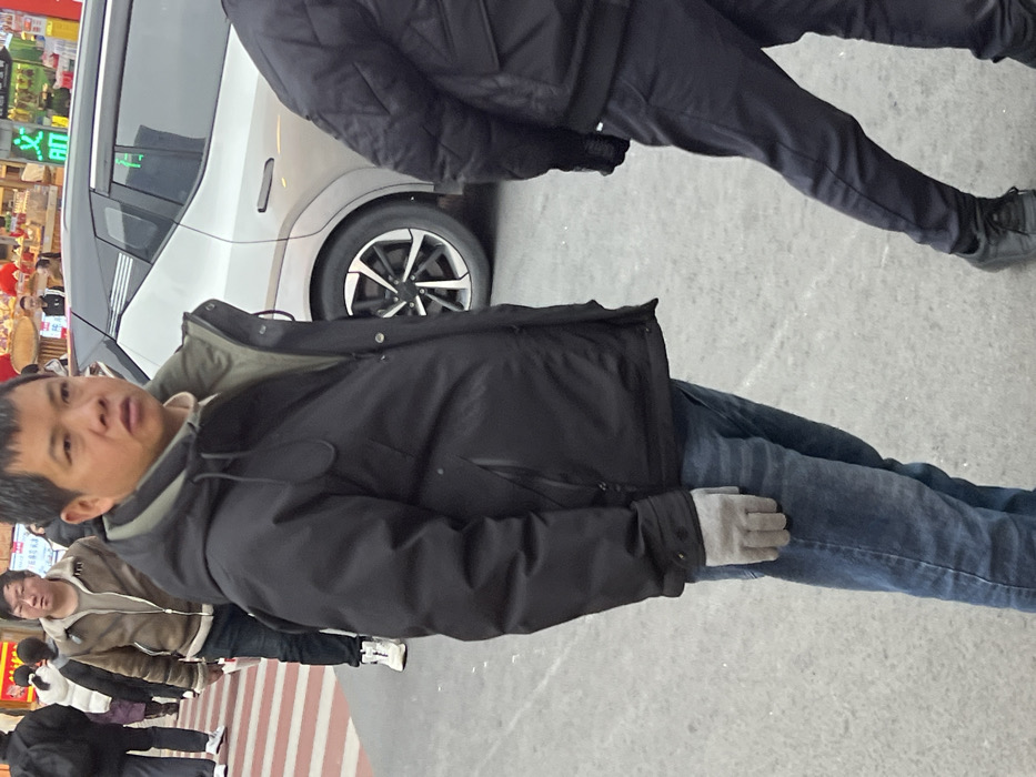 A street-level image depicts an urban scene in Yuzhong District, China. In the foreground, an adult male, of East Asian descent, stands facing slightly right of the camera with a neutral expression. He wears a black hooded puffer jacket with a visible olive green lining, a light cream-colored shirt underneath, blue denim jeans, and light grey gloves on his left hand.
To his immediate right, partially visible in the right foreground, is another individual from the waist down, wearing a black quilted jacket, dark trousers, and dark shoes, walking away from the camera. Behind the primary subject, a light-colored (possibly silver) SUV or crossover vehicle is parked or stationary, with its rear passenger side visible, showcasing a black roofline and a multi-spoke wheel design. A green character or symbol is adhered to its rear passenger window.
Further in the background, to the left of the primary subject, a red and white striped pedestrian crosswalk is visible on a grey paved surface. Multiple pedestrians are present, including a male figure in a brown jacket, dark jeans, and white shoes, walking across the crosswalk from left to right. Commercial establishments with illuminated signs bearing Chinese characters, along with red lanterns, are discernible in the distant background, indicating a bustling urban environment. The ground surface is grey pavement.